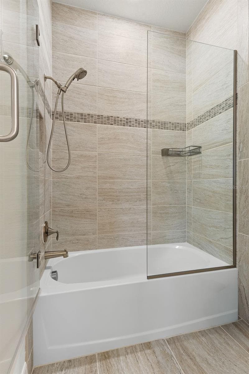 At the center of the Jack-and-Jill bath, the shared tub and shower are surrounded by floor-to-ceiling tile with a coordinating accent band, a glass enclosure, and a convenient handheld showerhead — connecting the two private vanity areas on either side. - 1528 FOREST GLEN CT, PALM HARBOR