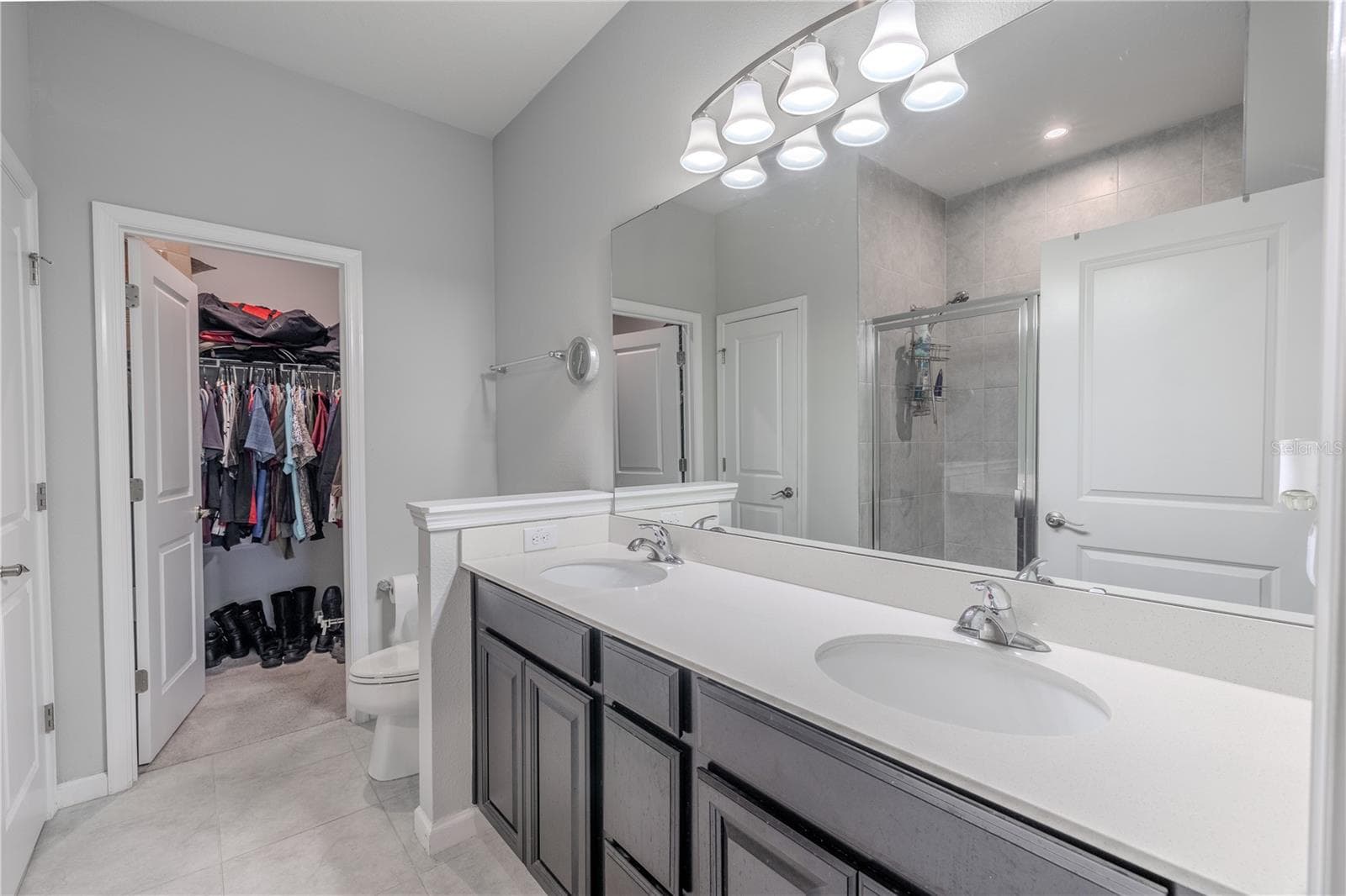 The primary ensuite features tile flooring, a long mirrored dual-sink vanity with generous storage and a bright downlight fixture, plus a glass-enclosed tiled shower and a second closet for added storage. - 3029 JACOB CROSSING LN, HOLIDAY