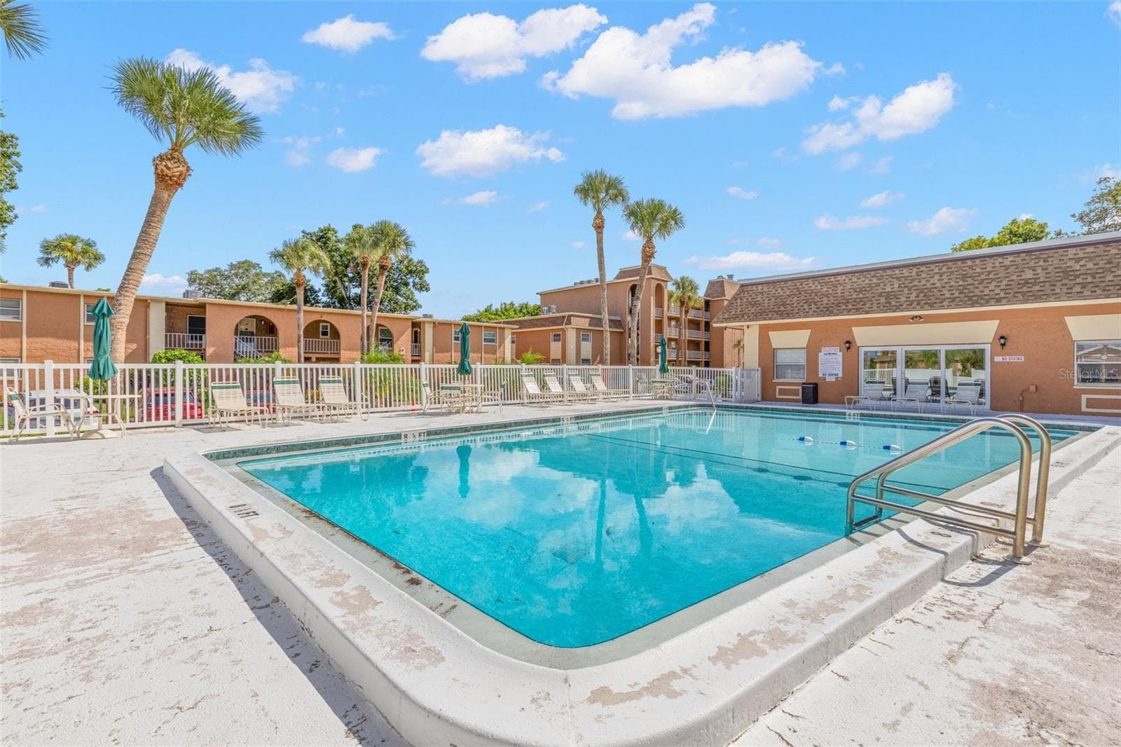 The community pool is surrounded by a spacious deck with lounge seating and umbrellas, while palms and the neighboring clubhouse create an inviting spot to relax outdoors. - 12760 INDIAN ROCKS RD #107, LARGO