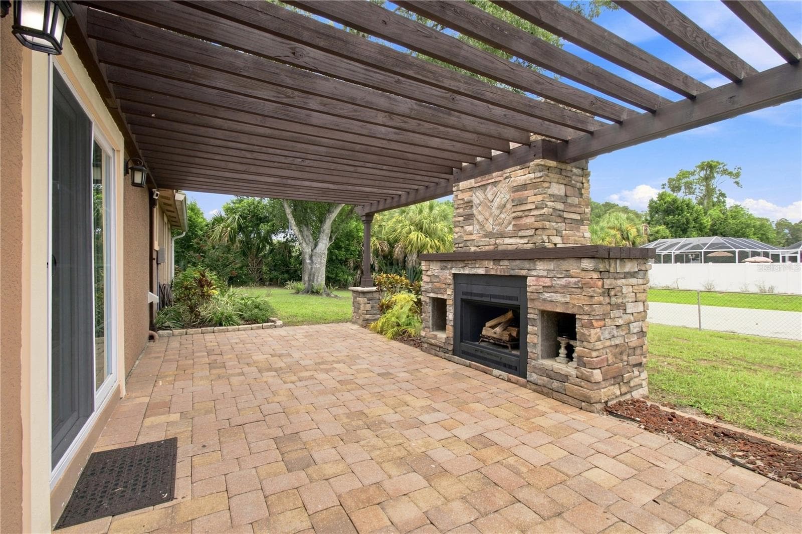 Crafted outdoor patio space featuring a large, rustic stone wood burning fireplace built under a wooden pergola and warm color pavers. - 9020 AUBURN WAY, TAMPA