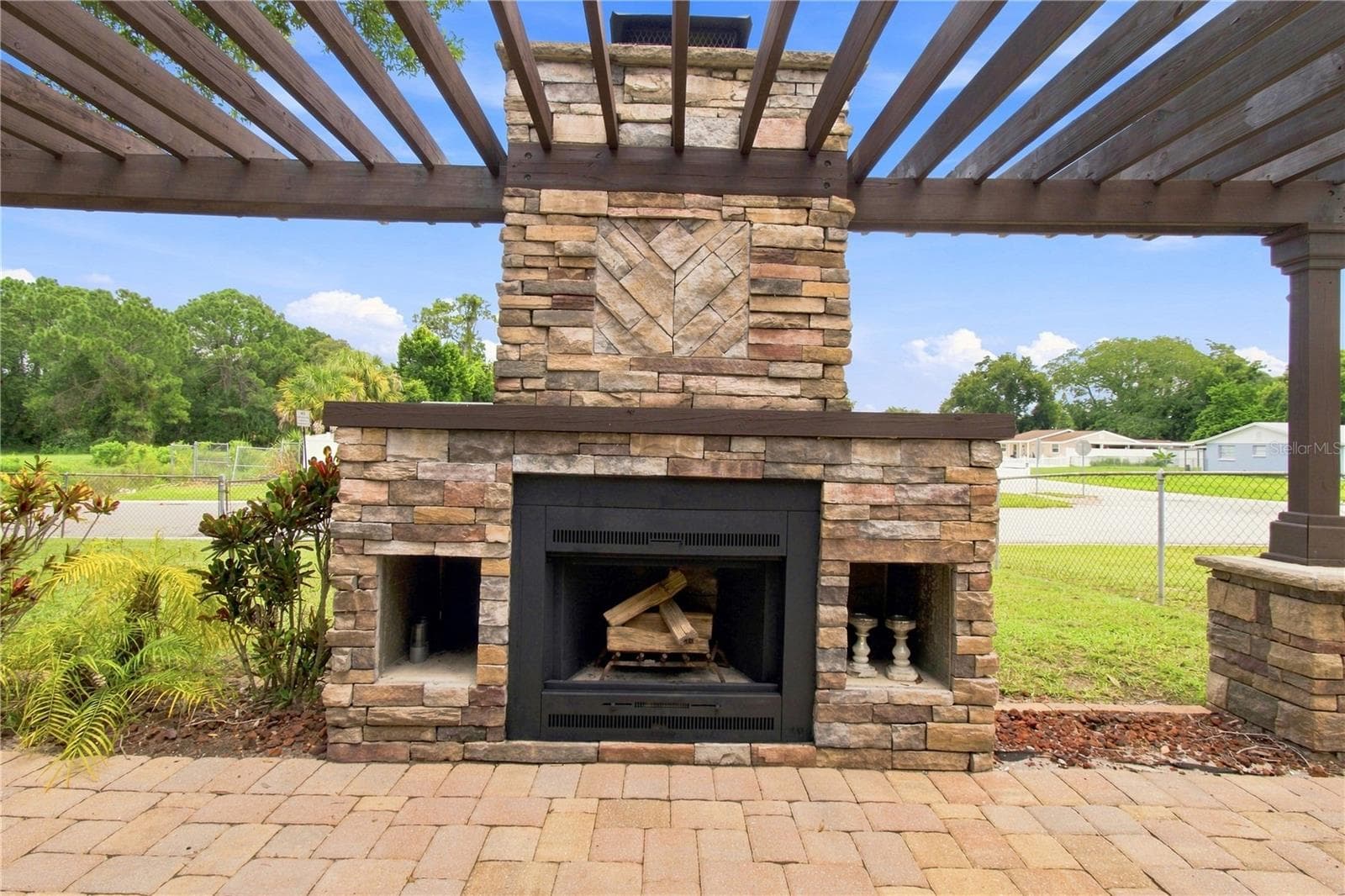 Crafted outdoor patio space featuring a large, rustic stone wood burning fireplace built under a wooden pergola and warm color pavers. - 9020 AUBURN WAY, TAMPA