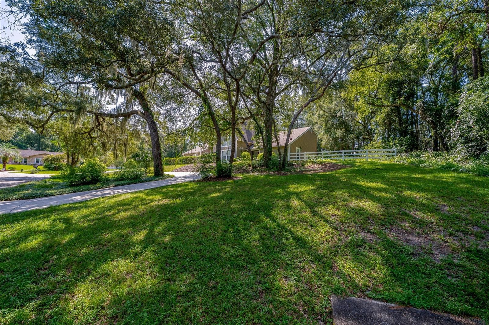 Expansive front lawn set beneath mature shade trees with a paved driveway and white fencing along the property line. - 2081 PENNSYLVANIA DR, DELAND