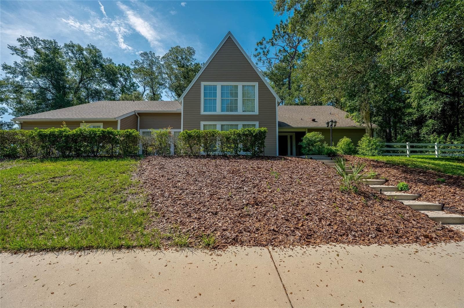 Front elevation of the two-story home featuring a steep roofline, tan vinyl siding, terraced landscaping steps, and lush surrounding greenery. - 2081 PENNSYLVANIA DR, DELAND