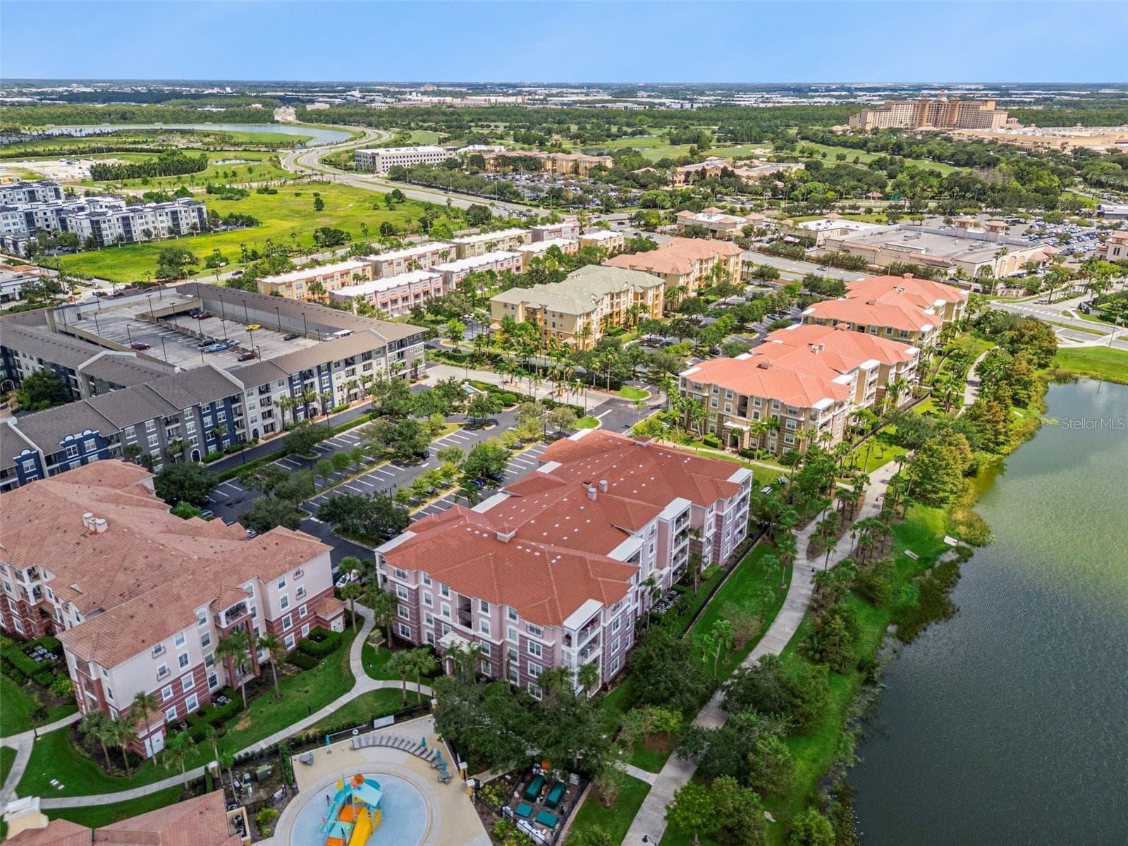 Lakefront Community Aerial:Expansive aerial shot showcasing tile-roofed condo buildings along the lake, paved walking trail, surface parking, and surrounding resort area. - 4840 CAYVIEW AVE #405, ORLANDO