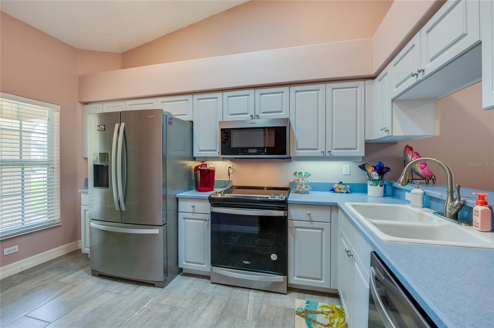 Functional kitchen equipped with stainless steel appliances, white cabinetry, and blue countertops. A large window brings in natural light, while high vaulted ceilings add depth. - 1413 COCONUT PALM CIR, PORT ORANGE