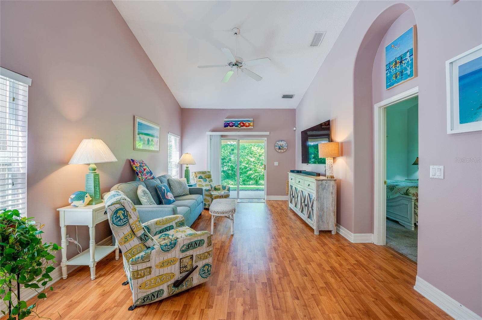 Spacious living room boasting dramatic vaulted ceilings, a central ceiling fan, and warm laminate flooring. Large sliding glass doors open directly to the outdoor patio space, while an arched doorway provides access to an adjoining bedroom. - 1413 COCONUT PALM CIR, PORT ORANGE