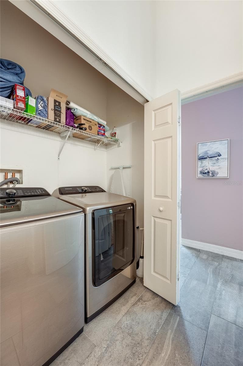 Dedicated laundry alcove equipped with side-by-side stainless steel appliances and overhead wire storage shelving. Grey tile flooring connects the utility space to the adjacent living areas. - 1413 COCONUT PALM CIR, PORT ORANGE