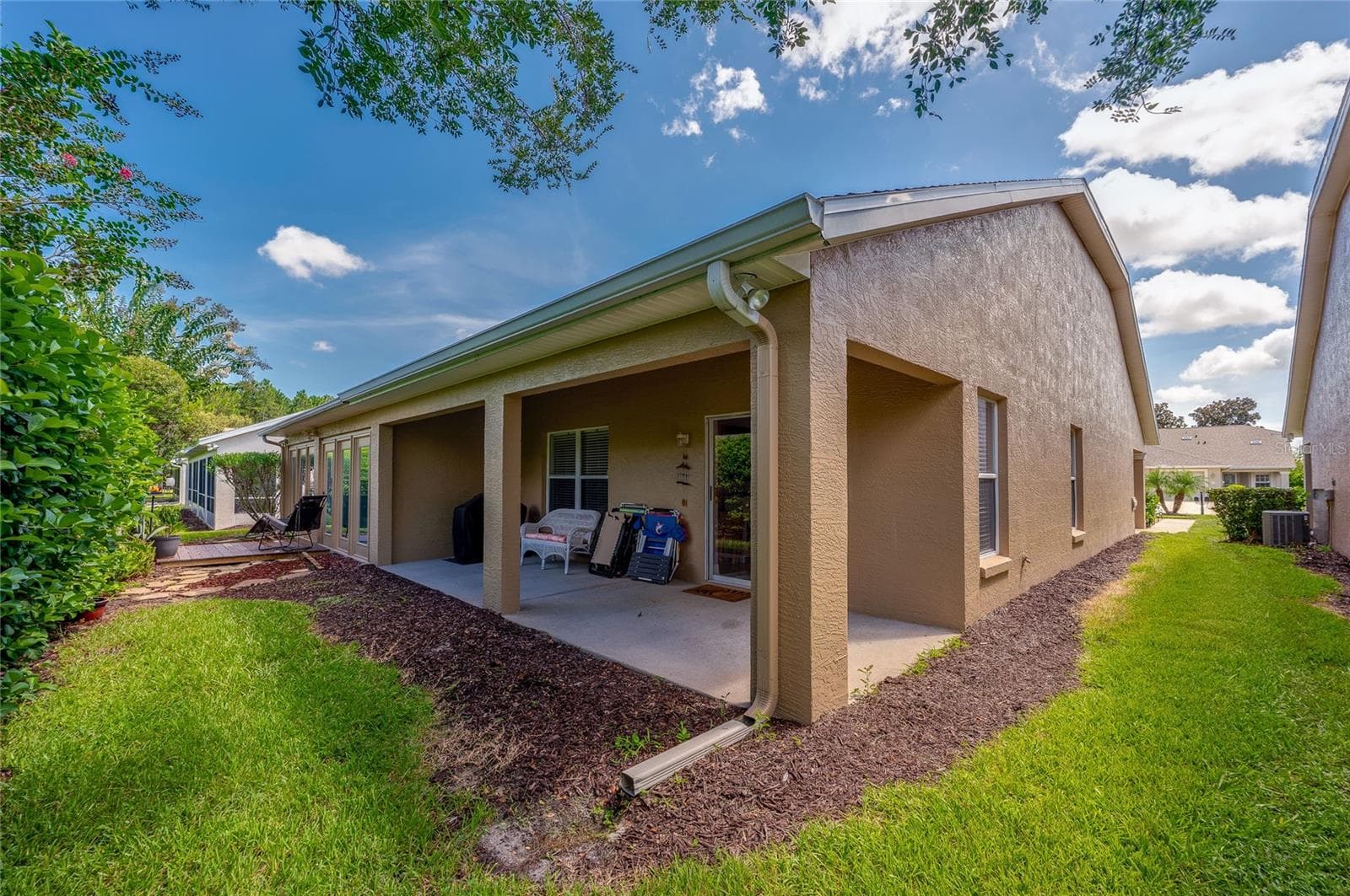 Exterior view of the rear elevation featuring a long covered patio and a well-maintained lawn bordered by mulch beds. Crisp stucco finishes and integrated roof gutters complete the exterior structure under clear blue skies. - 1413 COCONUT PALM CIR, PORT ORANGE