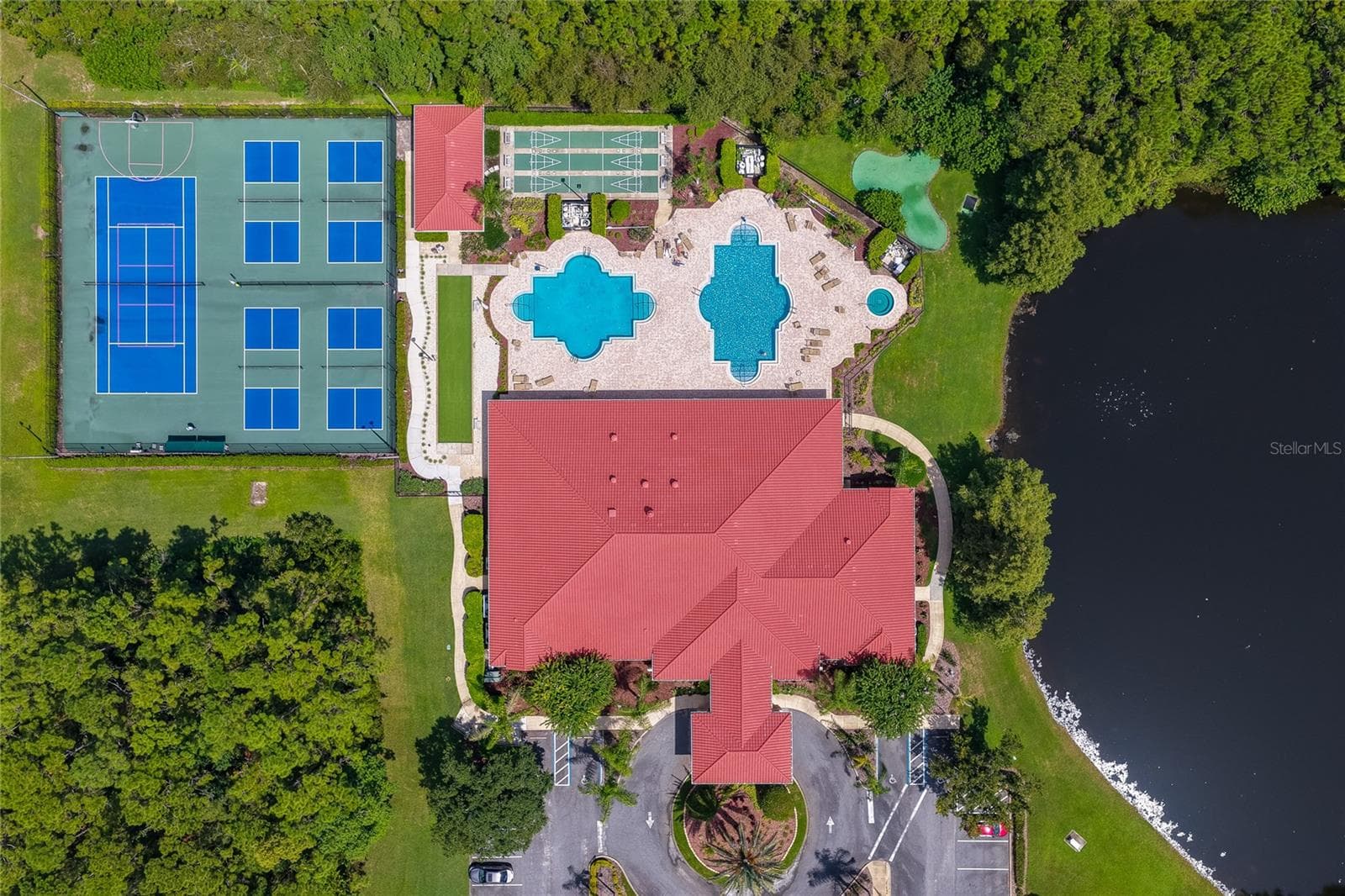 Top-down aerial view of community amenities including a spacious clubhouse with a distinct red roof, dual swimming pools, shuffleboard courts, and tennis courts. The facility is framed by a serene body of water and dense natural greenery. - 1413 COCONUT PALM CIR, PORT ORANGE