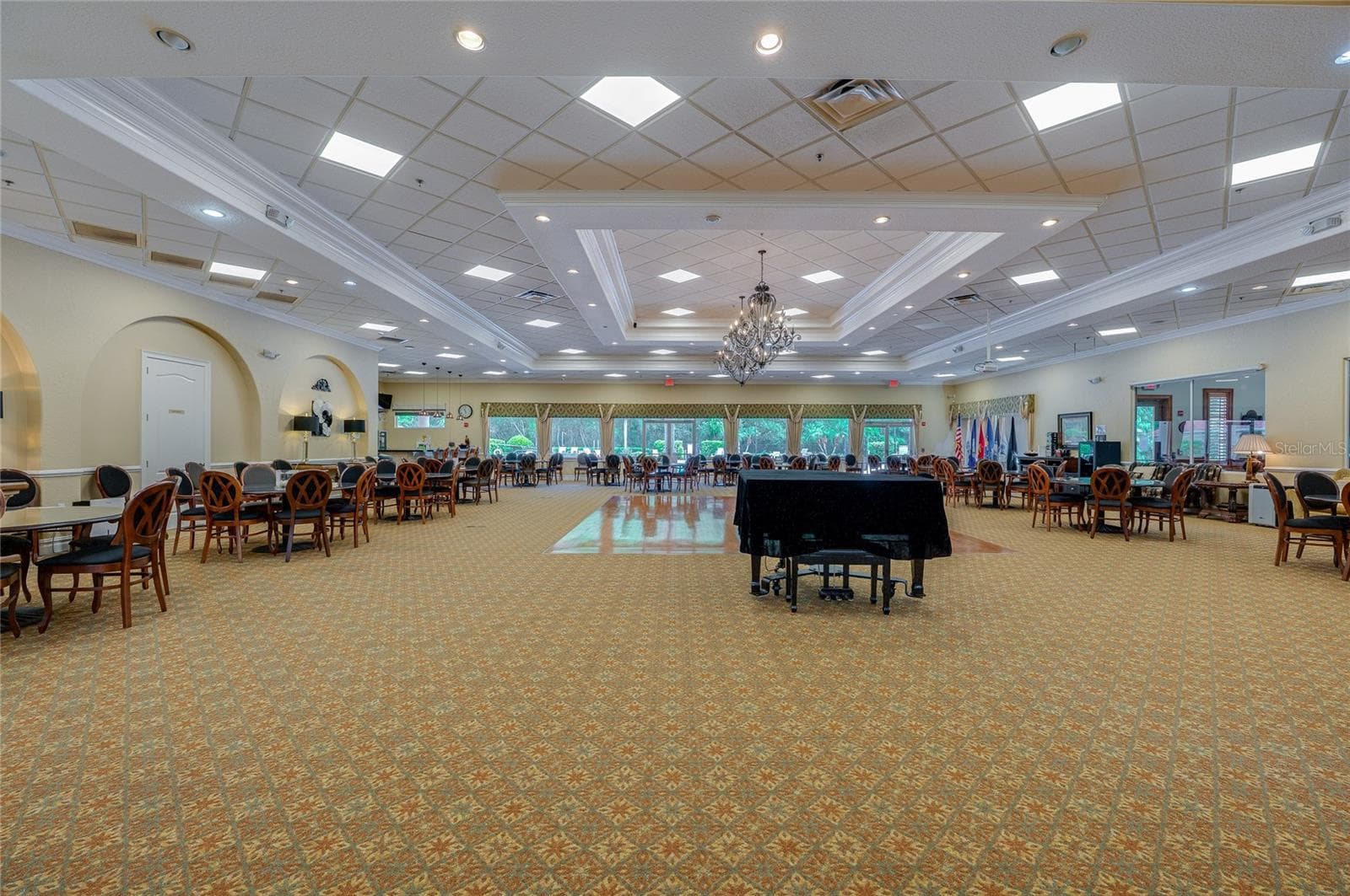 Grand event hall and dining room featuring drop tray ceilings, ornate chandelier lighting, patterned carpeting, and arched wall niches. Large glass doors along the far wall look out toward lush green landscaping. - 1413 COCONUT PALM CIR, PORT ORANGE