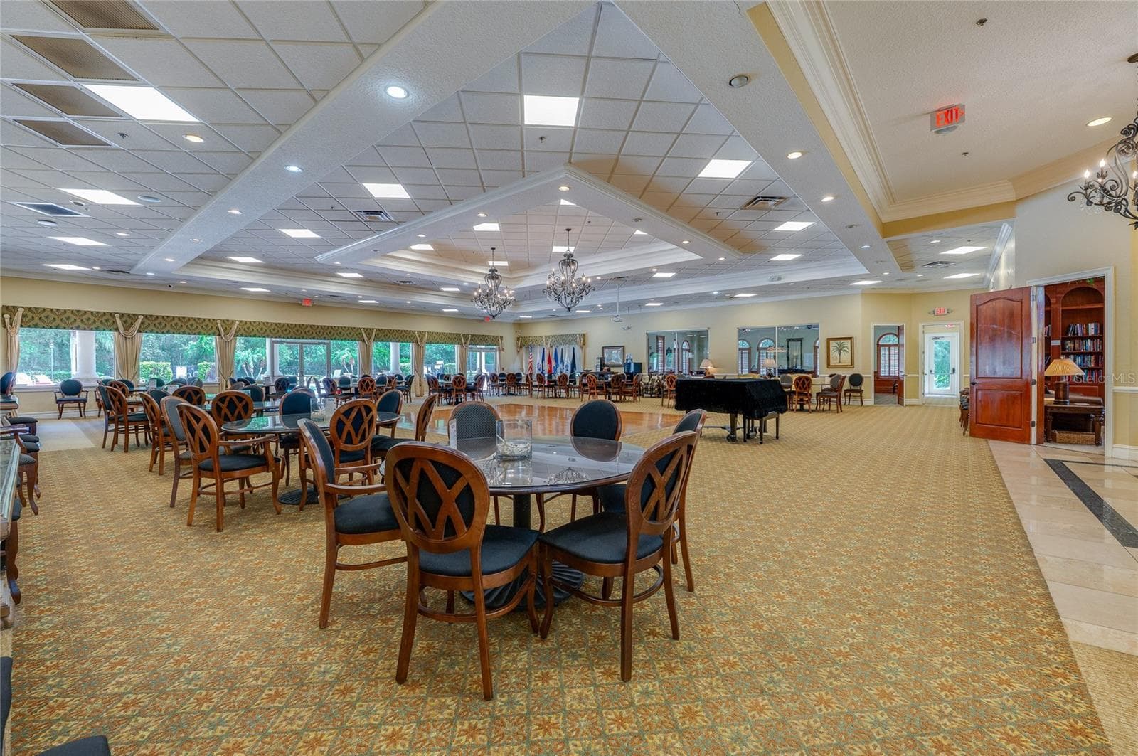 Expansive multi-purpose ballroom showcasing continuous patterned carpeting, multi-tiered tray ceilings with chandeliers, and a central polished dance floor. An open doorway on the side provides direct access to a quiet adjoining library space. - 1413 COCONUT PALM CIR, PORT ORANGE
