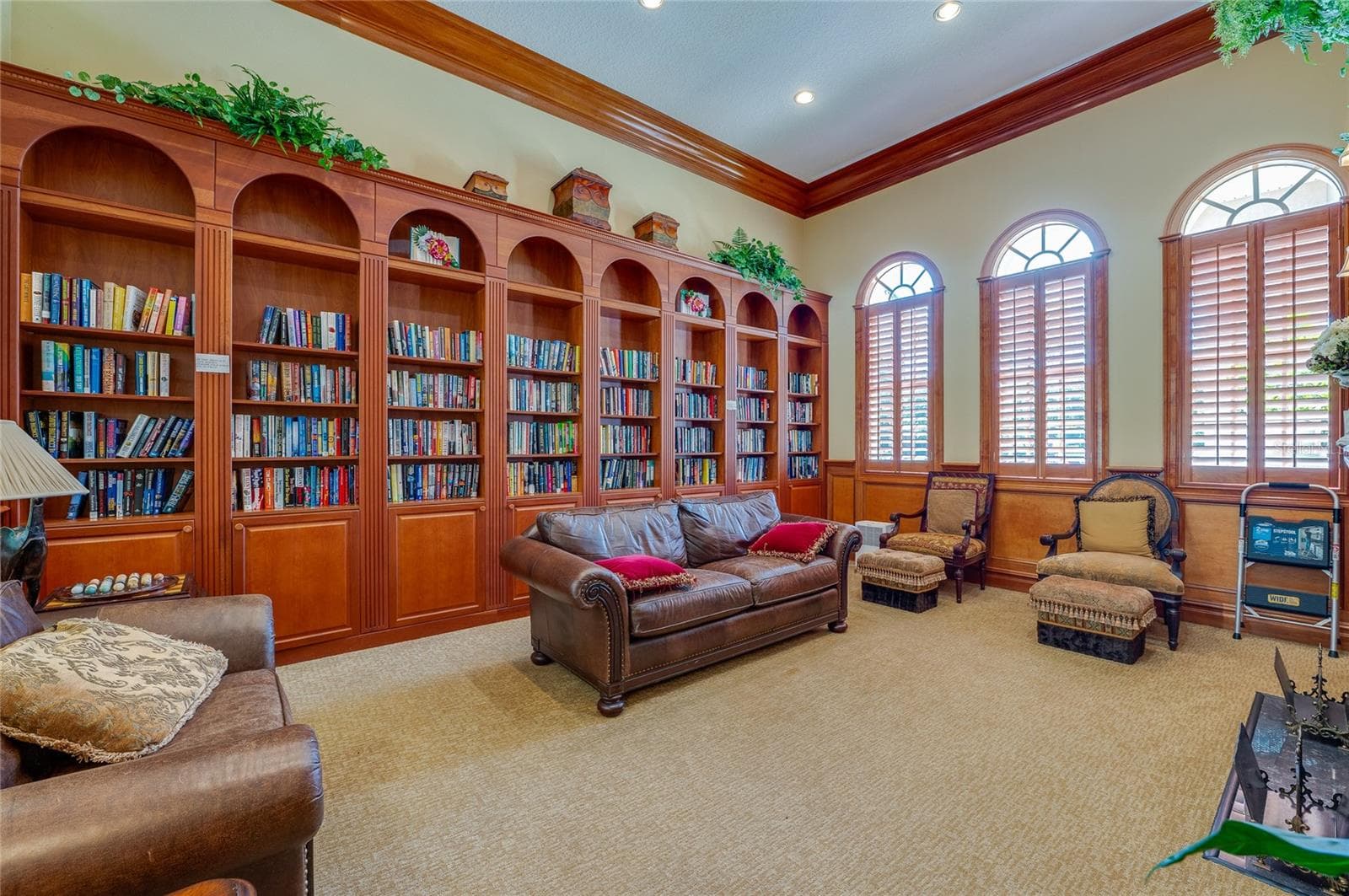 Cozy reading library framed by floor-to-ceiling rich wood built-in bookcases, warm crown molding, and neutral carpeting. Three large arched windows outfitted with wooden plantation shutters bring soft natural light into the space. - 1413 COCONUT PALM CIR, PORT ORANGE