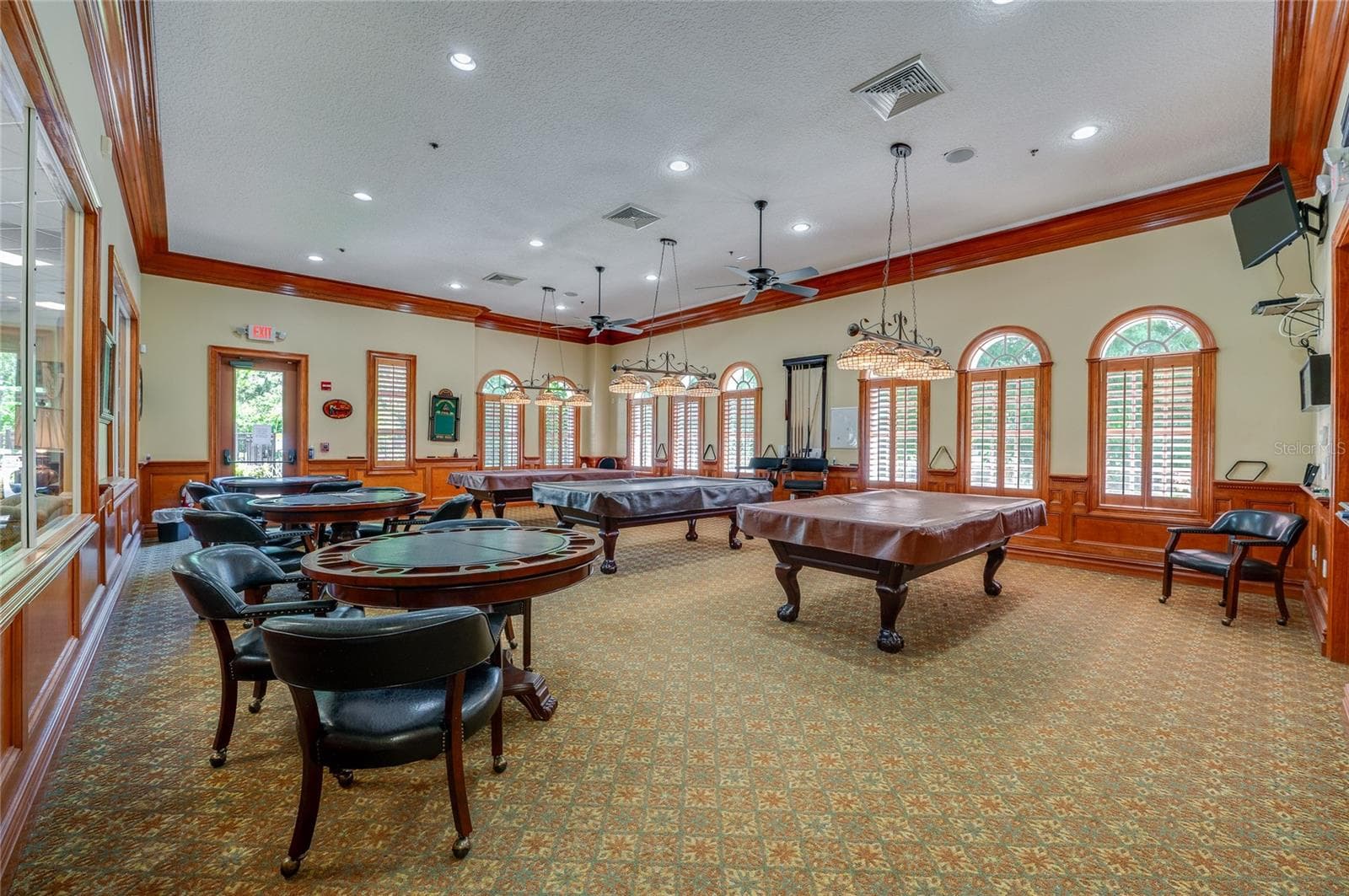 Spacious game and billiards room featuring recessed lighting, warm wood crown molding, and wainscoting throughout. Tall arched windows with wooden shutters illuminate the room, which connects to adjacent common spaces via large interior viewing windows. - 1413 COCONUT PALM CIR, PORT ORANGE