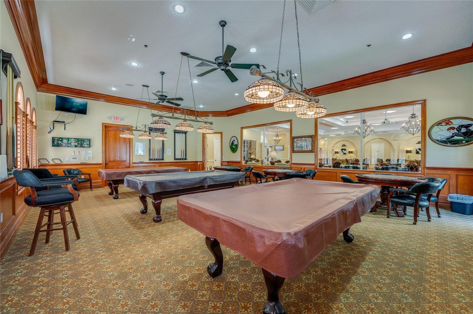 Secondary angle of the game room showcasing detailed wood wall trim, hanging stained-glass pendant lights, and overhead ceiling fans. An interior glass partition window provides a clear view into the main hall. - 1413 COCONUT PALM CIR, PORT ORANGE