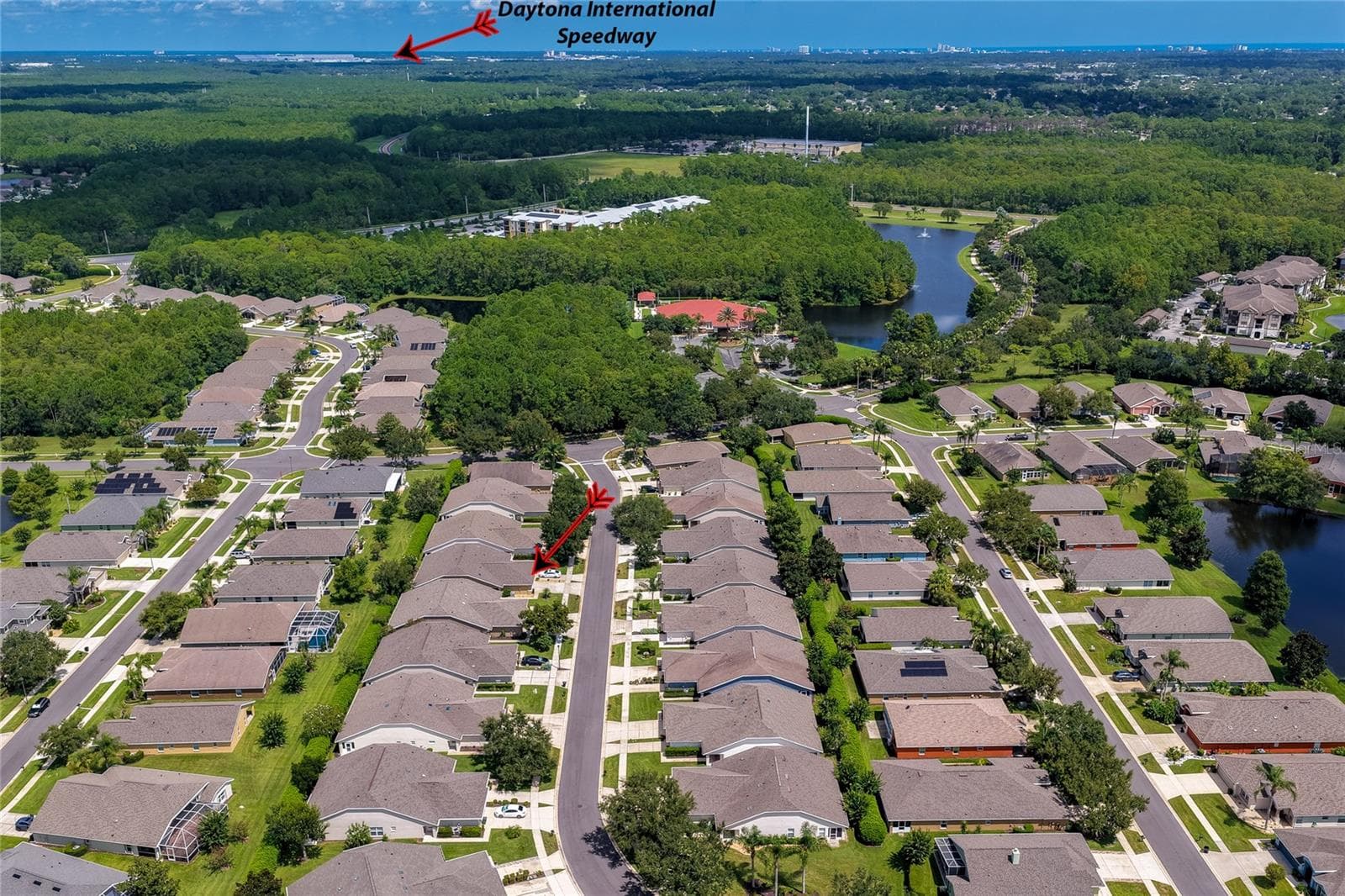 High-altitude aerial view showcasing the expansive residential development surrounded by dense woodlands, with a directional arrow pointing toward the iconic Daytona International Speedway in the distance. - 1413 COCONUT PALM CIR, PORT ORANGE