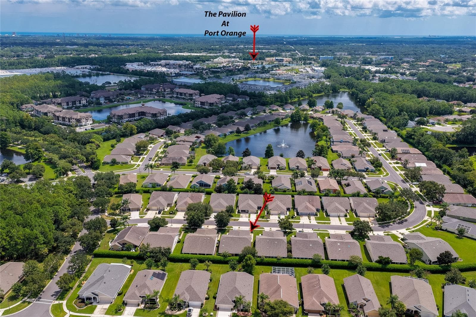 Broad aerial view capturing the residential neighborhood layout, surrounding water features, and green foliage, with a red arrow pointing toward the nearby Pavilion at Port Orange shopping area. - 1413 COCONUT PALM CIR, PORT ORANGE