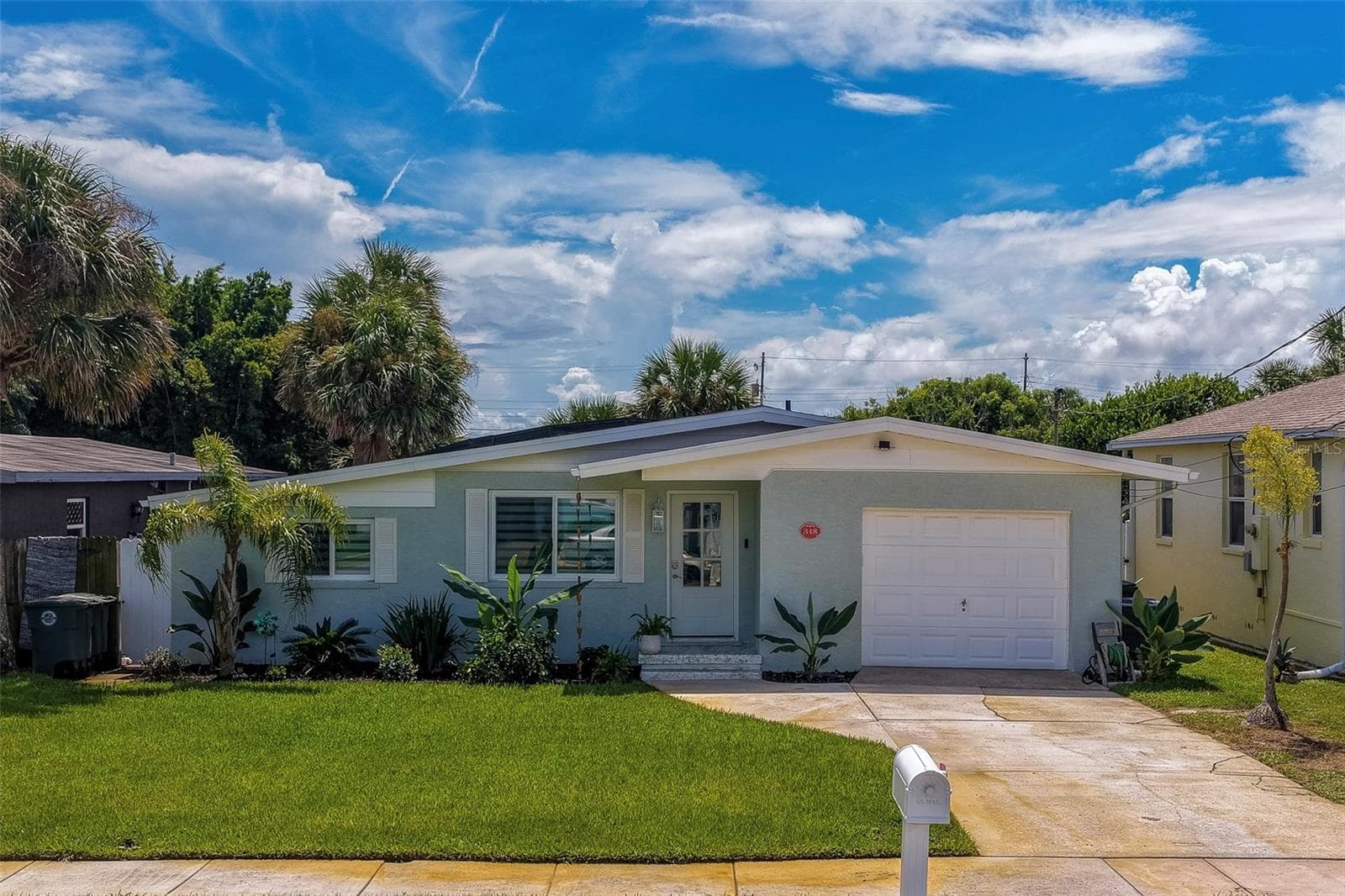 Charming single-story exterior showcasing a bright lawn, lush tropical landscaping, and a clean driveway leading to an attached garage under a vibrant blue sky. - 348 WOODLAND AVE, DAYTONA BEACH
