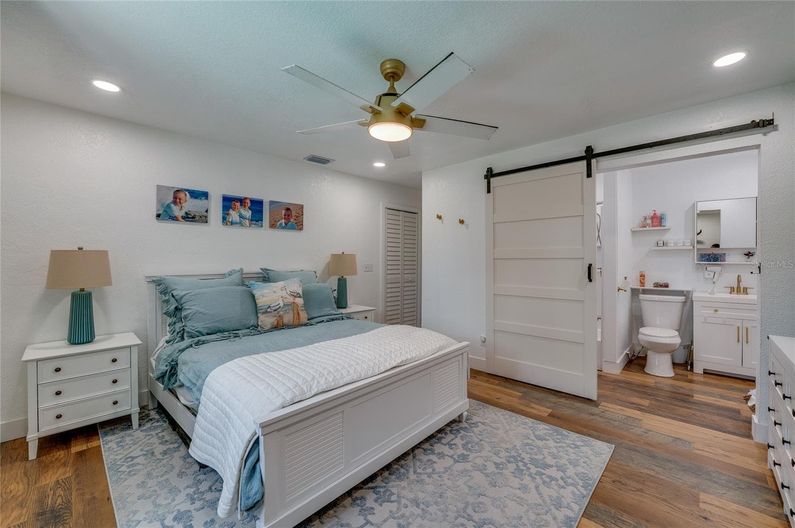 Spacious primary bedroom suite showcasing warm LVP flooring, a ceiling fan, and direct access to an ensuite bathroom through a sliding white barn door. - 348 WOODLAND AVE, DAYTONA BEACH