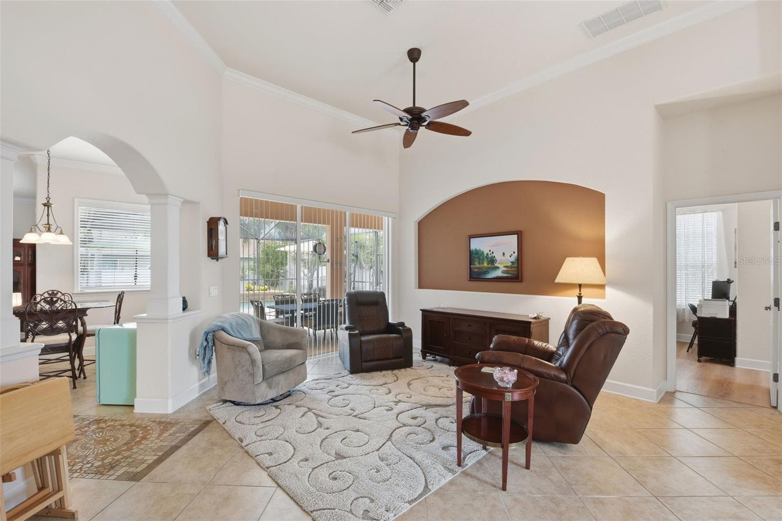 Living Room – Spacious living area with high ceilings, crown molding, a ceiling fan, and architectural archways that create an open connection to adjoining spaces. Sliding glass doors bring in natural light and provide access to the outdoor Lanai and Pool. - 114 VIOLA DR, AUBURNDALE