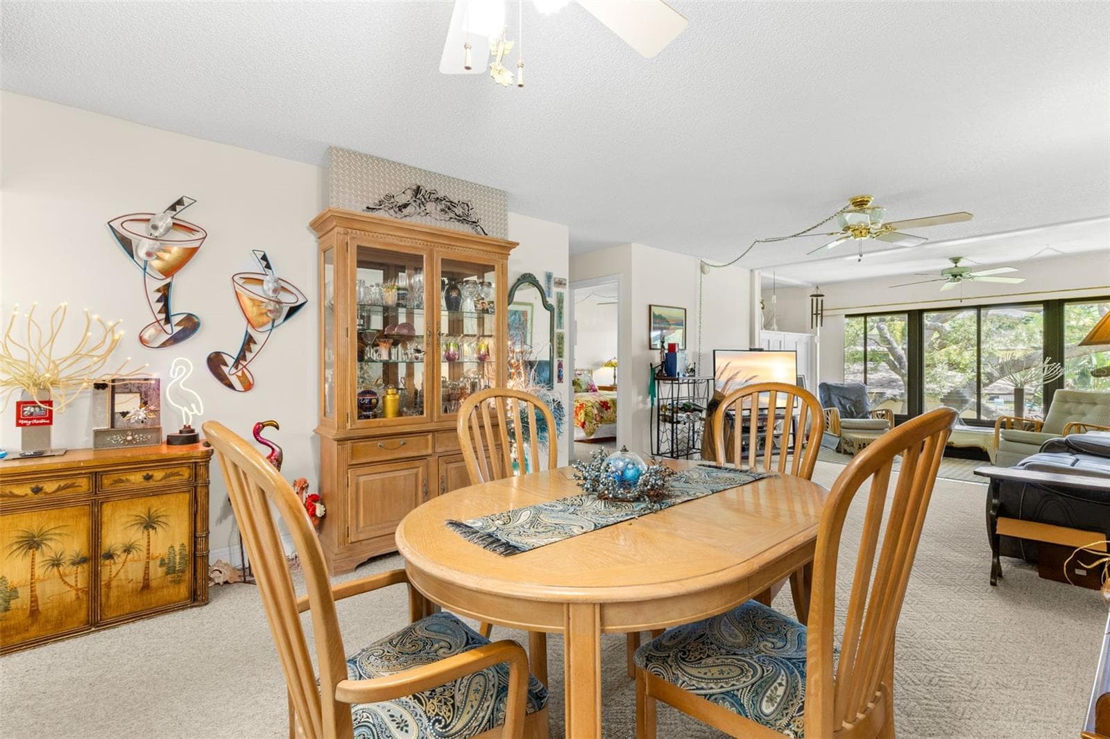 From the dining area, the sightline extends through the living room to the wall of windows at the far end. Neutral carpeting continues through both spaces, with ceiling fans overhead and the bedroom hall tucked off to the side. - 12300 VONN RD #3204, LARGO
