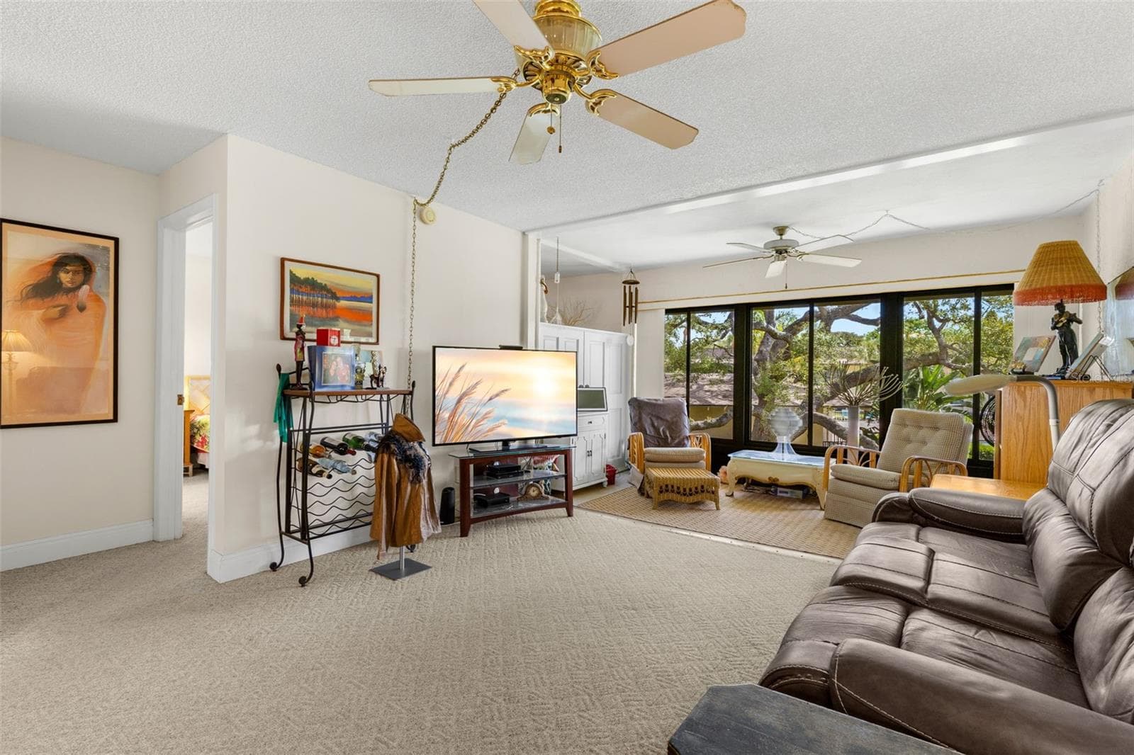 The living room opens directly into the enclosed Florida room, creating a second sitting area framed by a broad wall of windows. Neutral carpeting continues through both spaces, while ceiling fans add comfort overhead. - 12300 VONN RD #3204, LARGO