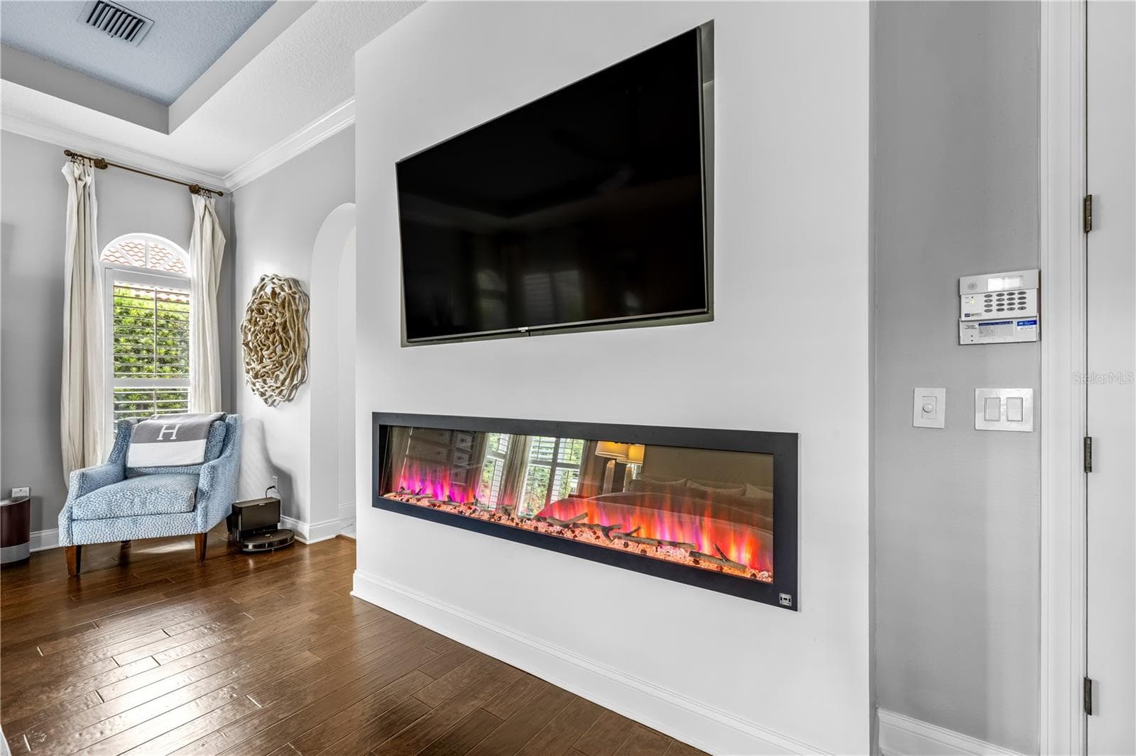 Linear electric fireplace beneath a mounted TV with an arched window and plantation shutters. - 1921 FLORESTA VIEW DR, TAMPA
