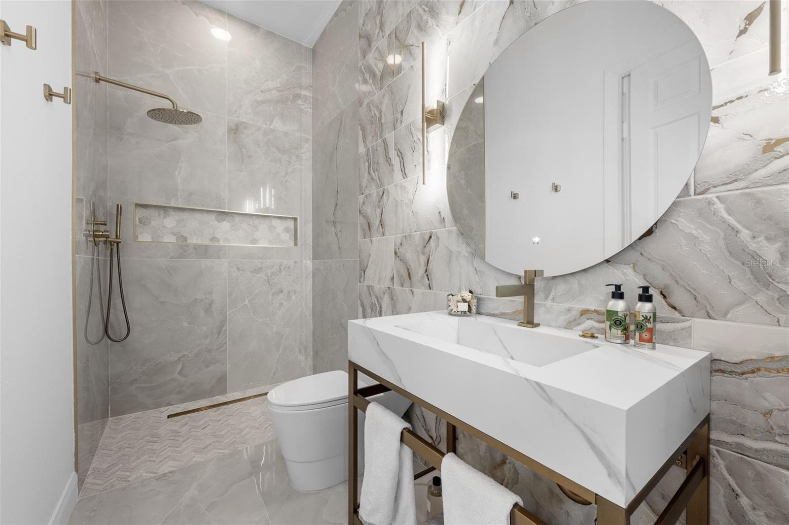 3rd Bathroom - Marble-look tile walls, console sink with brass frame, round mirror, and walk-in shower with brass fixtures. - 1921 FLORESTA VIEW DR, TAMPA