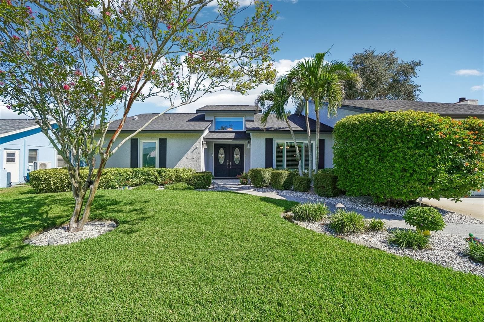 Front Exterior – Mature palms, a flowering tree, lush lawn, and neatly shaped shrubs create an inviting setting along the home’s front elevation. - 971 CORTLAND WAY, PALM HARBOR