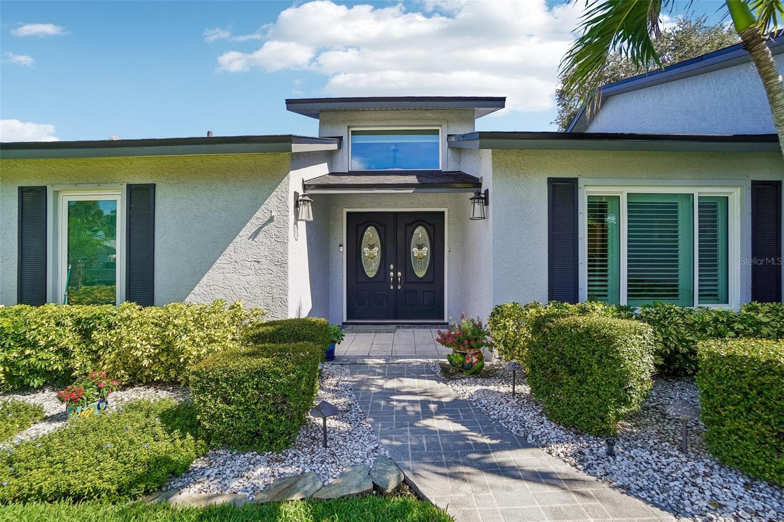 Front Entry – A paved walkway surrounded by manicured landscaping leads to decorative double entry doors, accented by exterior lighting and a large window above. - 971 CORTLAND WAY, PALM HARBOR