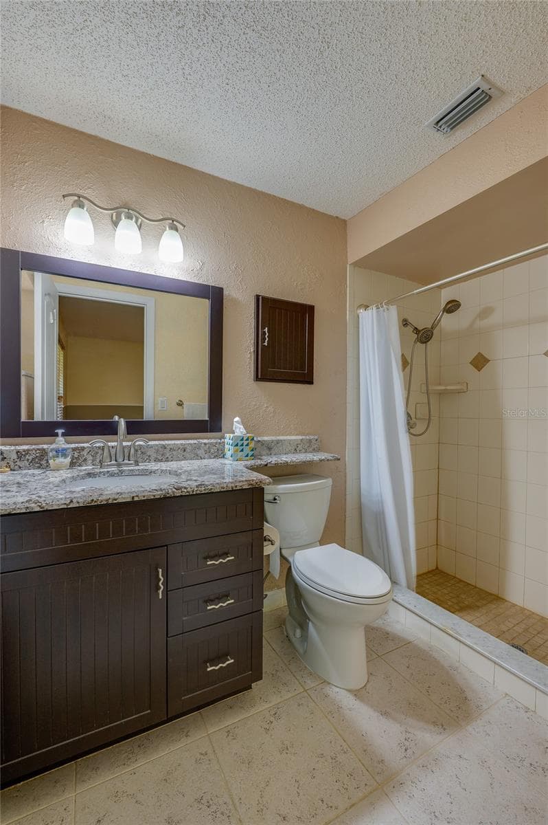 Primary bathroom showcasing a dark wood vanity with granite countertops, tiled flooring, a wide framed mirror, and a walk-in tiled shower. - 4669 SECRET RIVER TRL, PORT ORANGE