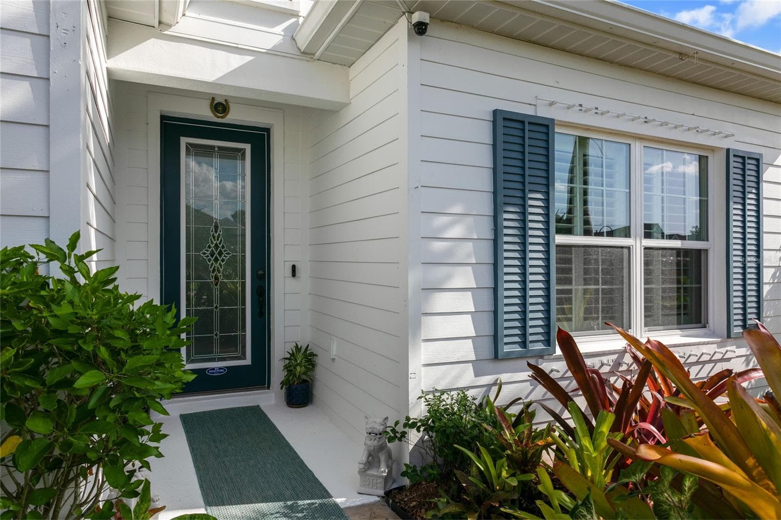 Inviting front entry featuring a teal designer door with decorative glass panels, complemented by crisp white siding and blue shutters. Tropical landscaping with vibrant foliage enhances the curb appeal, creating a warm and welcoming first impression.The home’s entryway exudes charm and sophistication, highlighted by a striking teal door with intricate glass detailing and framed by clean white siding and coastal blue shutters. Lush tropical landscaping adds texture and color, creatinga beautifully curated welcome that reflects Florida's vibrant lifestyle. - 10625 FALLING LEAF CT, PARRISH