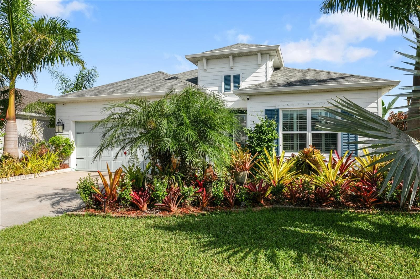 Beautiful front elevation featuring a light coastal façade, blue shutters, tropical landscaping, and a well-maintained lawn that enhances the home’s inviting curb appeal - 10625 FALLING LEAF CT, PARRISH