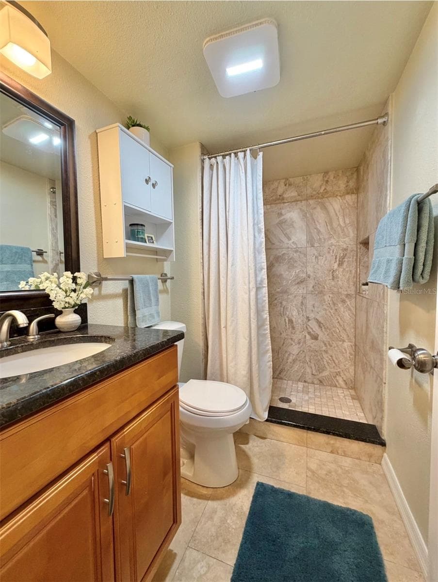The second bathroom features luxurious travertine floors, a floor-to-ceiling tiled walk-in shower with a niche, granite countertops, a storage vanity, new satin nickel fixtures, a storage cabinet above the toilet, and updated lighting. - 3815 N OAK DR #G71, TAMPA
