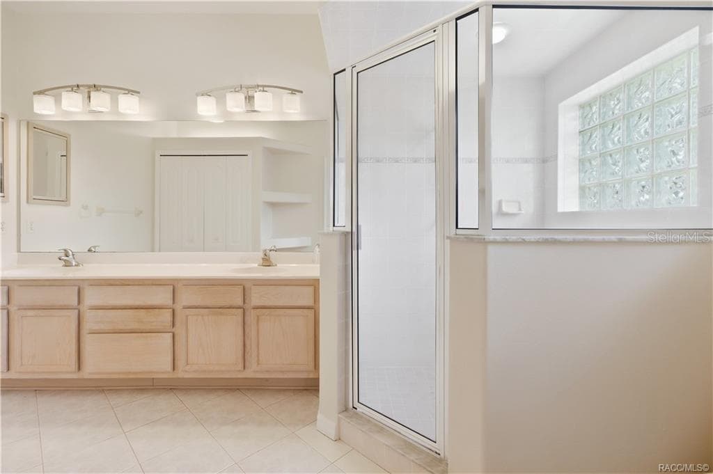 PRIMARY BATHROOM – Primary bath featuring a long dual-sink vanity, expansive wall mirror, tile flooring & large glass-enclosed walk-in shower accented by a glass-block window. - 6410 W CANNONDALE DR, CRYSTAL RIVER