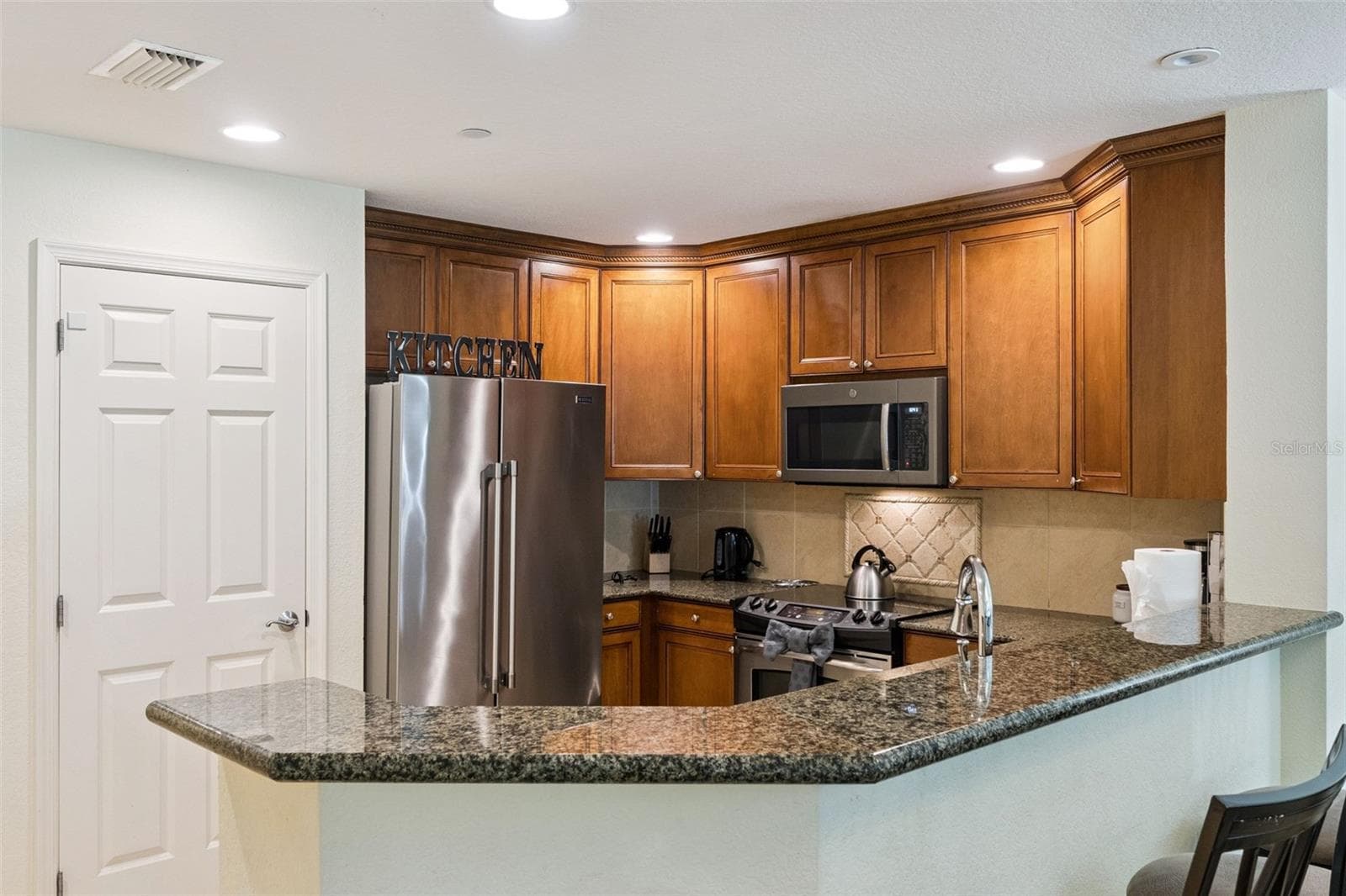 Kitchen Wrap-Around Bar: Angle highlighting the generous wrap-around granite counter space, deep wood upper and lower cabinetry, tile floor, and stainless steel appliances. - 7656 WHISPER WAY #102, REUNION