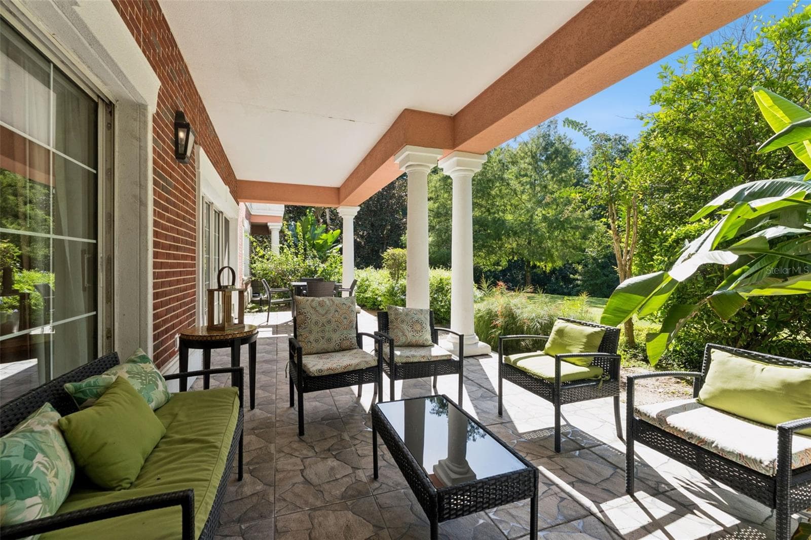 Covered Patio Seating: Covered outdoor patio with stone-patterned tile flooring, classic red brick wall accents, comfortable wicker lounge seating, and views of surrounding tropical flora. - 7656 WHISPER WAY #102, REUNION