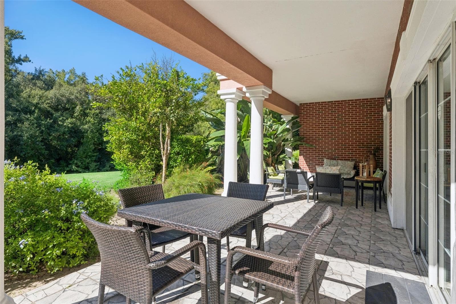Outdoor Dining Area: Spacious covered patio section featuring a four-person outdoor wicker dining set, white support columns, sliding glass door access, and lush greenery surroundings. - 7656 WHISPER WAY #102, REUNION
