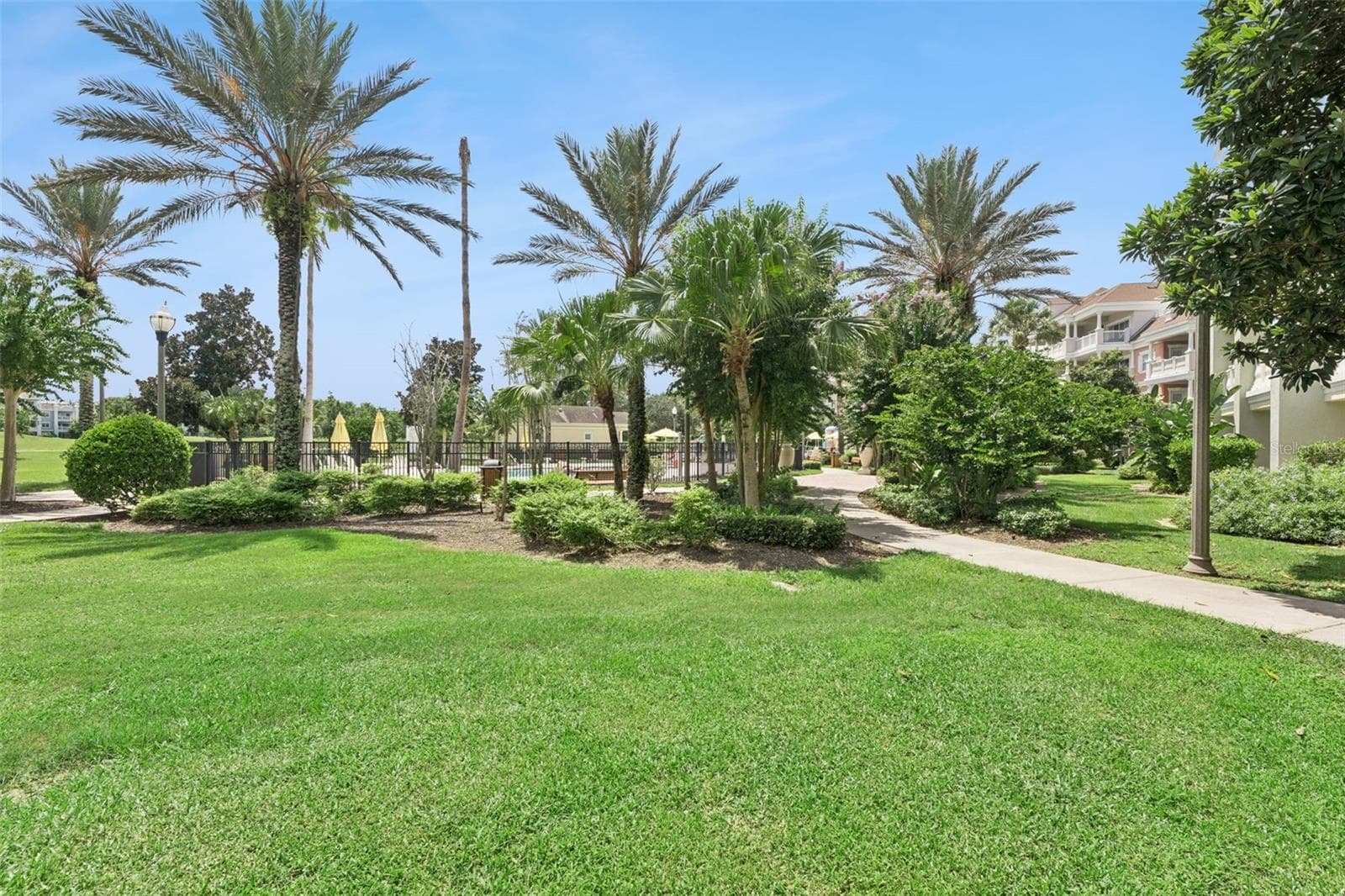 Landscaped Grounds & Courtyard: Ground-level view featuring manicured green lawns, paved walking paths, tropical palm trees, and gated access to the community pool facilities. - 7656 WHISPER WAY #102, REUNION