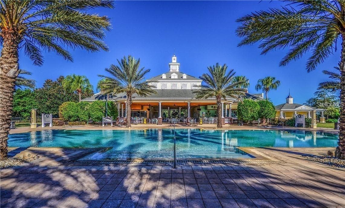 Resort Pool & Community Clubhouse: Sun-drenched resort swimming pool framed by tall palm trees, showcasing the grand community clubhouse and covered patio pavilion. - 7656 WHISPER WAY #102, REUNION