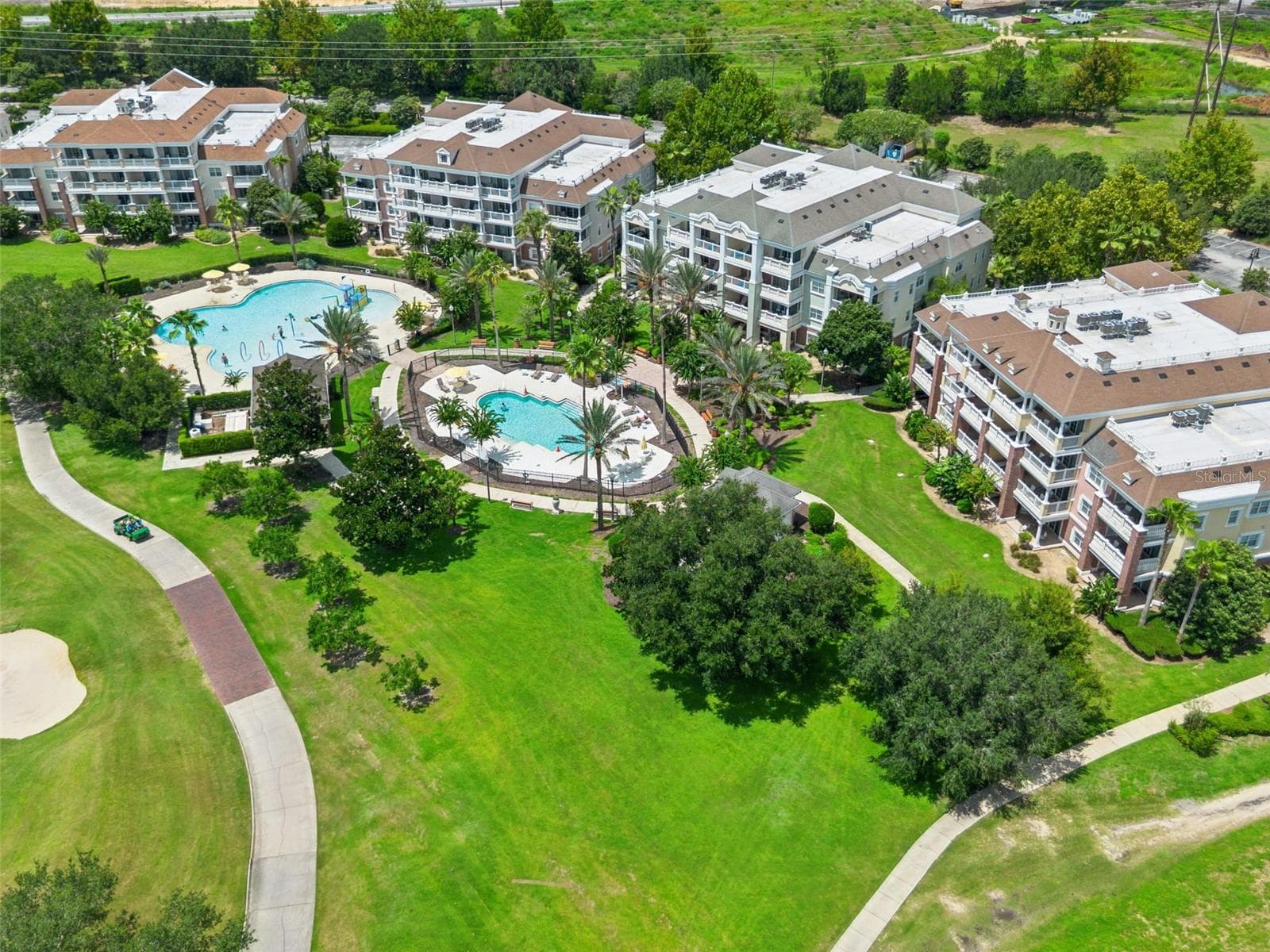 Pool Amenities & Fairway View: Elevated aerial shot highlighting the resort pool grounds, paved golf cart paths, expansive green lawns, and residential buildings. - 7656 WHISPER WAY #102, REUNION