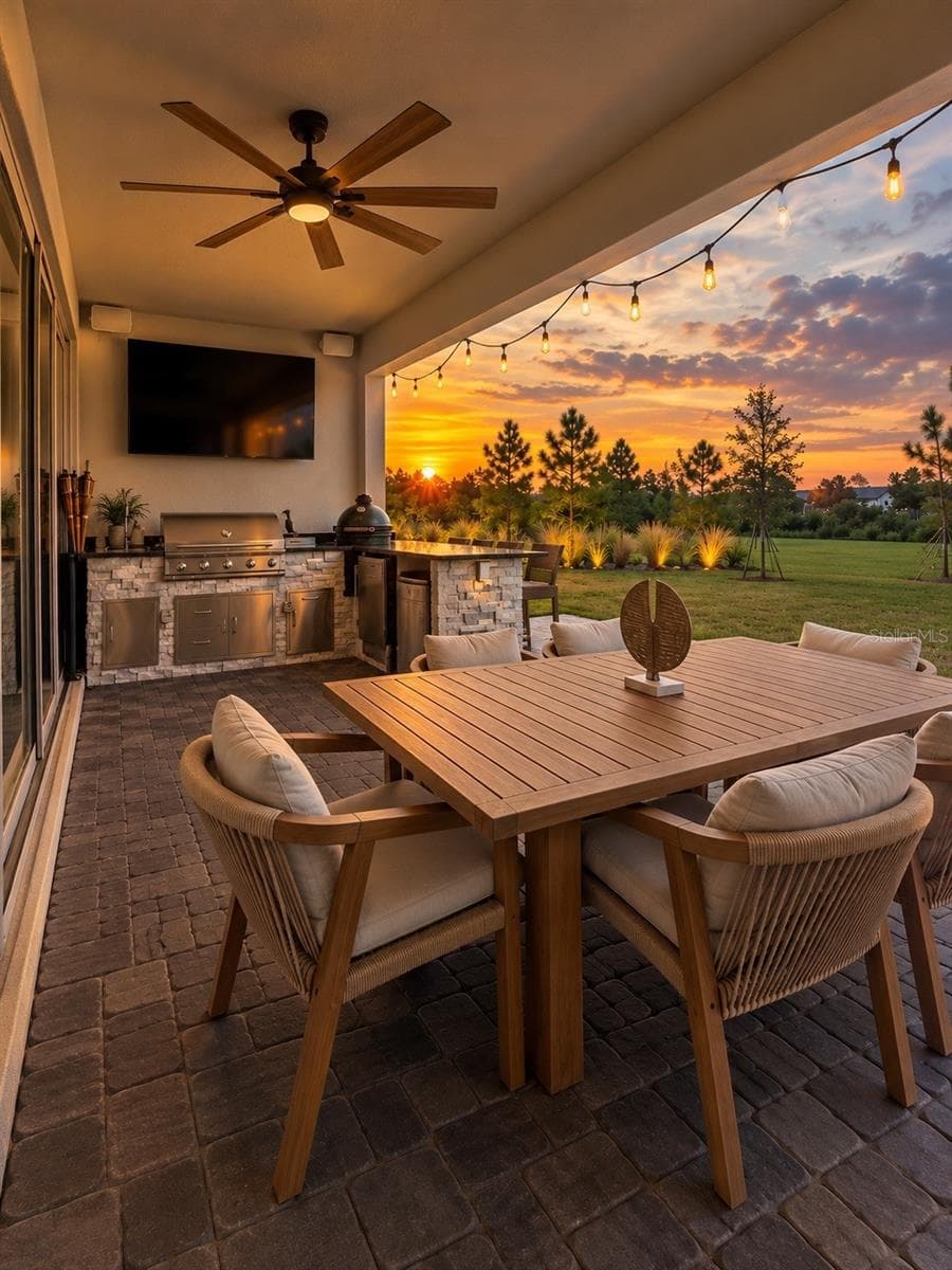 Covered outdoor living area with built-in summer kitchen, dining space, TV, ambient lighting, and peaceful sunset views over the landscaped green space. - 6735 REVERIE PARK AVE, ORLANDO