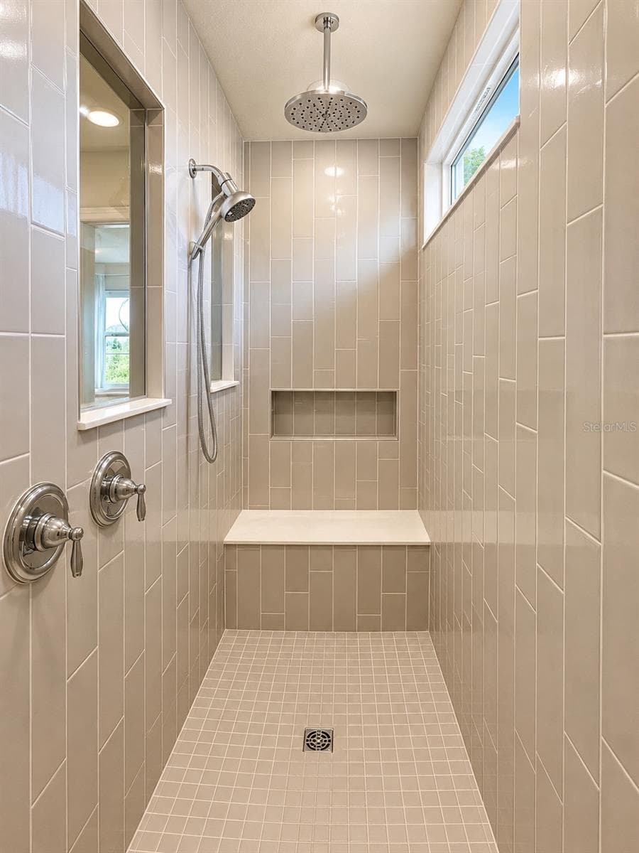 Spacious walk-in shower featuring floor-to-ceiling tile, built-in bench and niche, dual shower system with rainfall showerhead and handheld fixture, plus a window providing natural light - 6735 REVERIE PARK AVE, ORLANDO