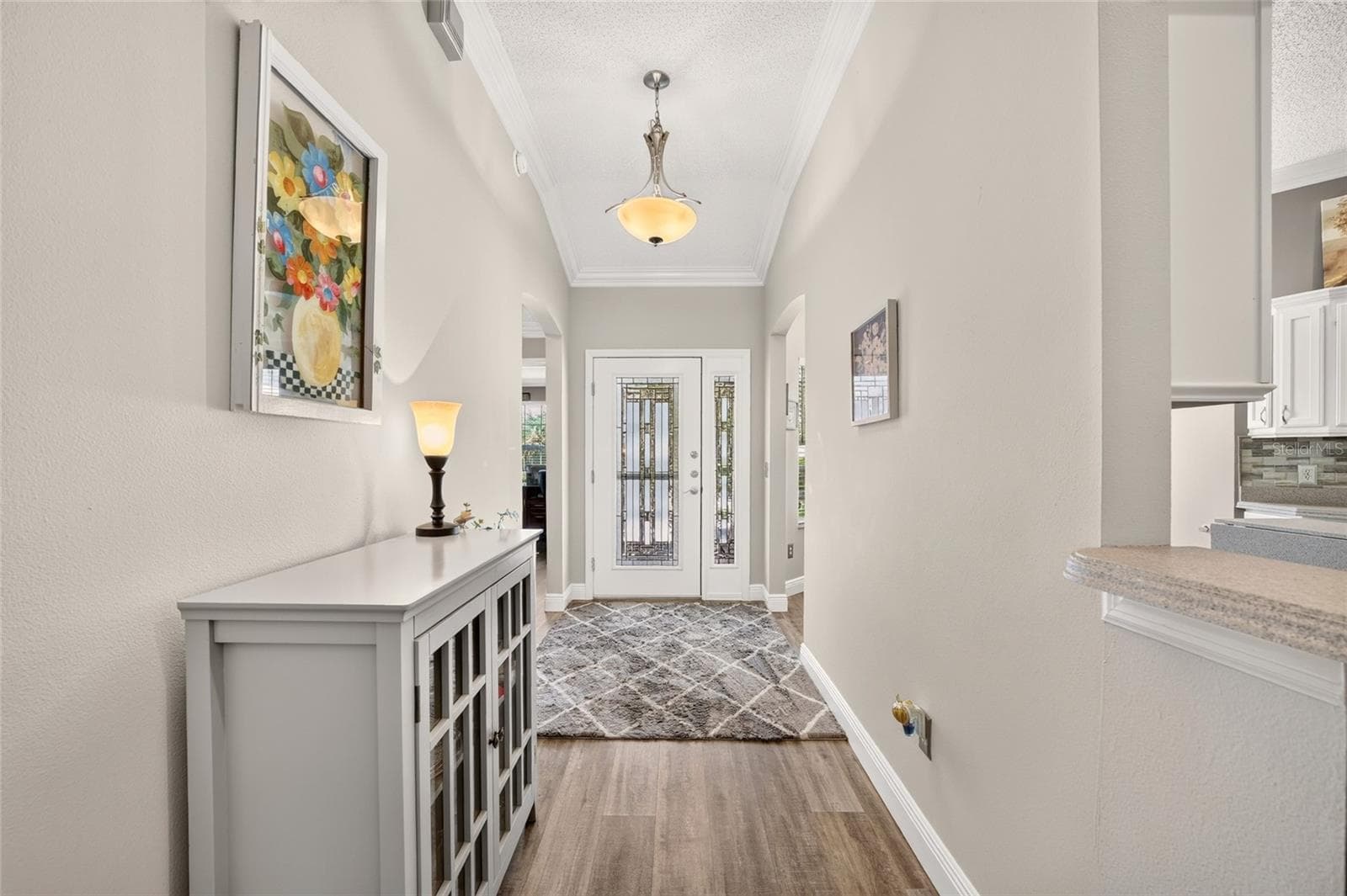 Foyer: Bright entry foyer features crown molding, wood-look flooring, a decorative glass front door with sidelight, and an open connection to the surrounding living spaces. - 2749 BANYAN PL, THE VILLAGES