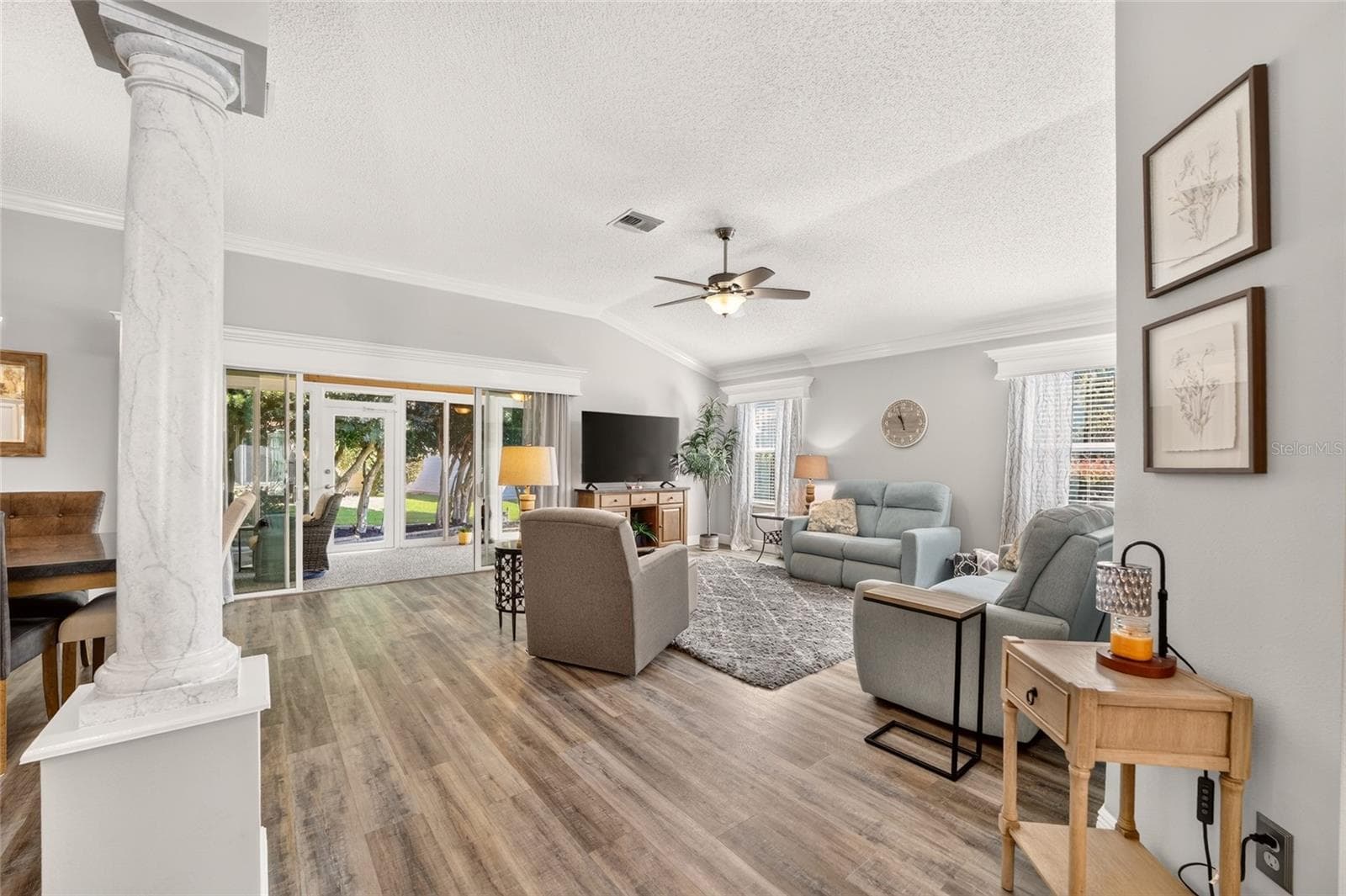 Living Room: Spacious living area offers wood-look flooring, crown molding, a ceiling fan, abundant natural light, and sliding glass doors extending the space toward the outdoors. - 2749 BANYAN PL, THE VILLAGES