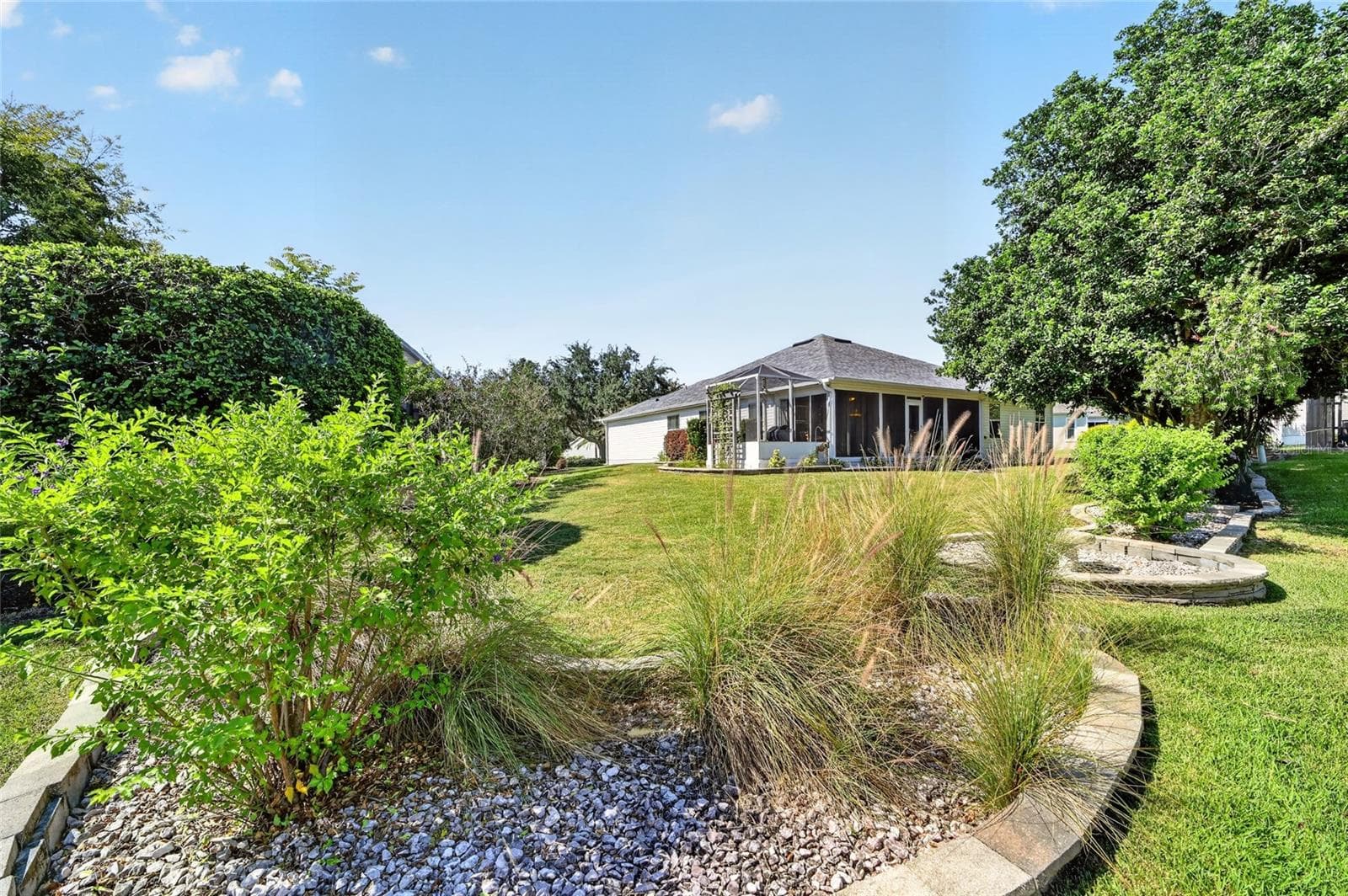 Backyard: Landscaped backyard features an open lawn, established plantings, decorative garden beds, mature trees, and views of the home's screened outdoor spaces. - 2749 BANYAN PL, THE VILLAGES