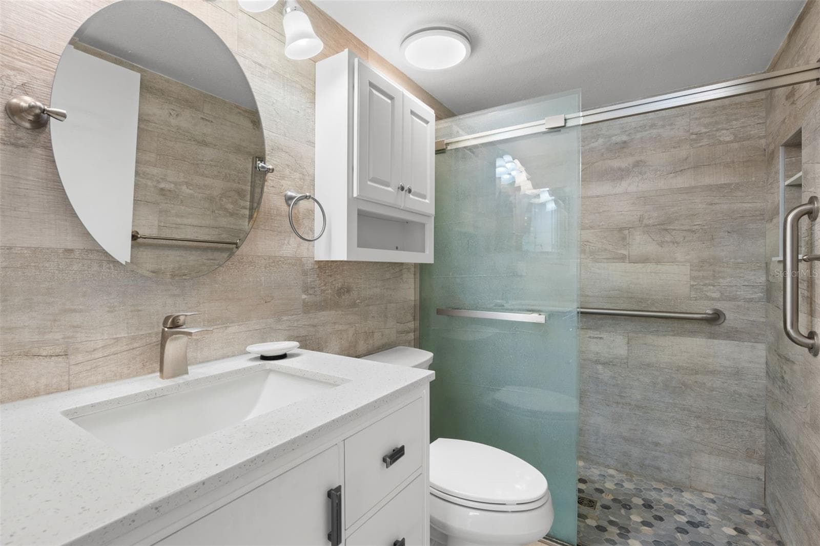 Updated in 2021, the primary ensuite features a sleek white vanity, large round mirror, and a walk-in shower with wood-look tile, pebble-style flooring, frameless glass, and grab bars. - 3188 LAKE PINE WAY S #F1, TARPON SPRINGS