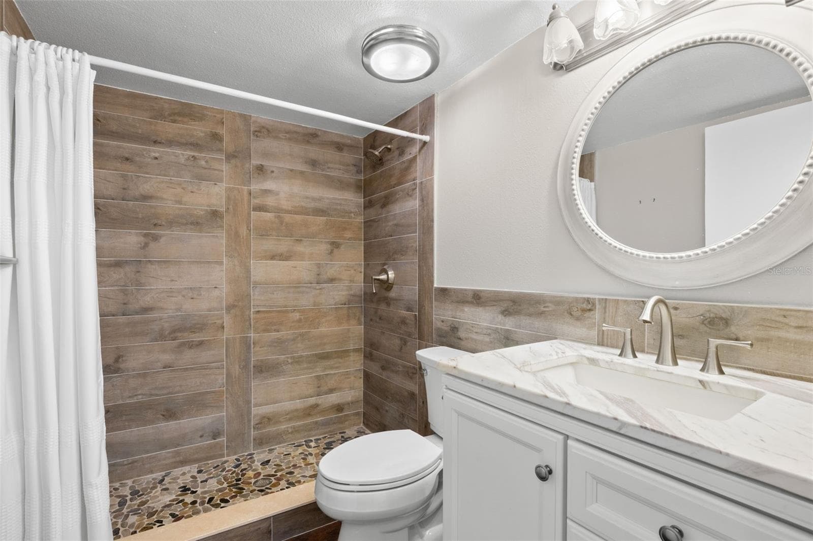 The updated second bath features a white vanity, oval mirror, and a tiled walk-in shower with wood-look walls and a pebble-style floor. - 3188 LAKE PINE WAY S #F1, TARPON SPRINGS