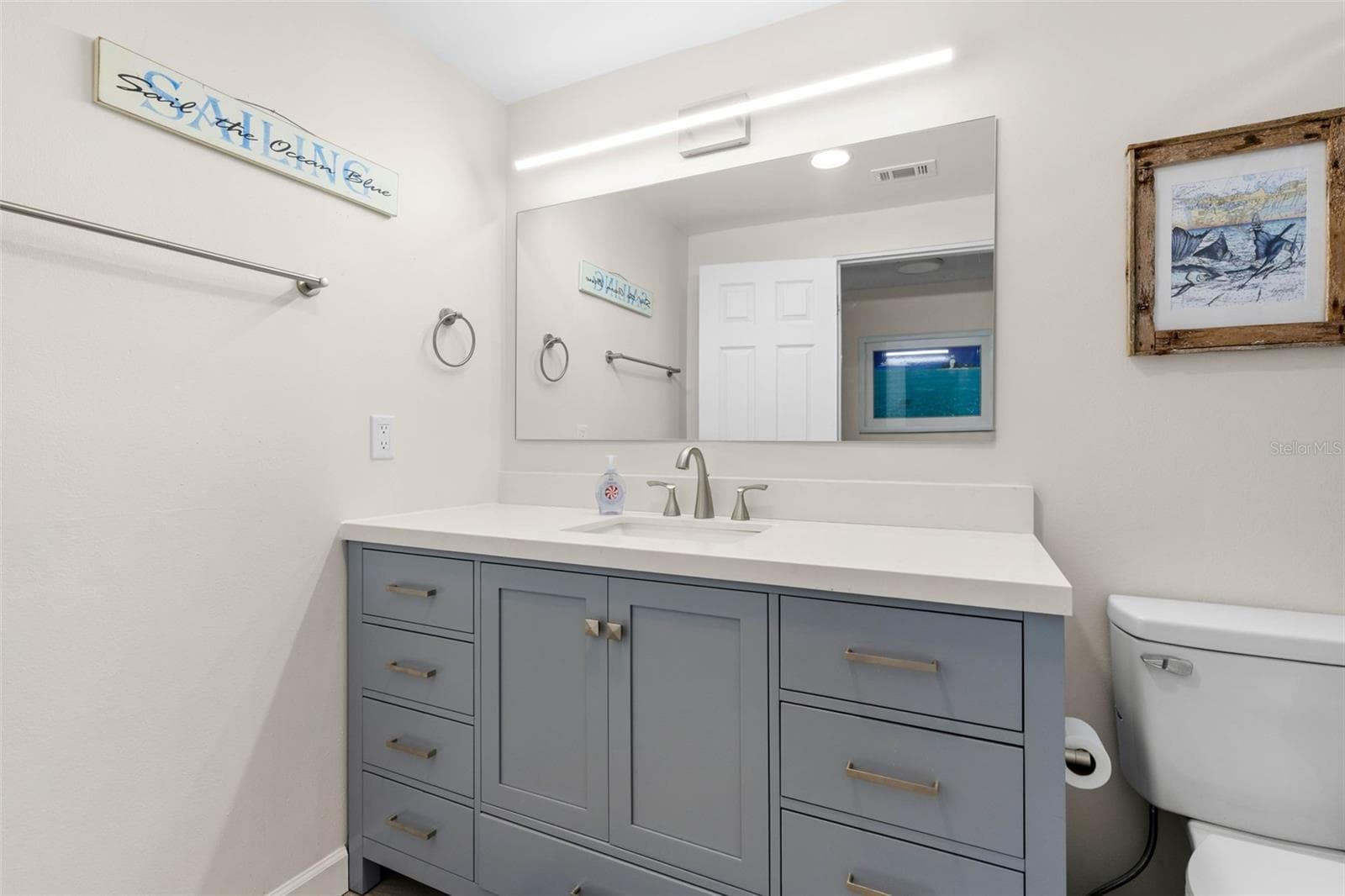The remodeled primary bath (2025) features a wide gray vanity with abundant drawer storage, a sleek white countertop, oversized mirror, and contemporary linear lighting. - 332 WESTWINDS DR, PALM HARBOR
