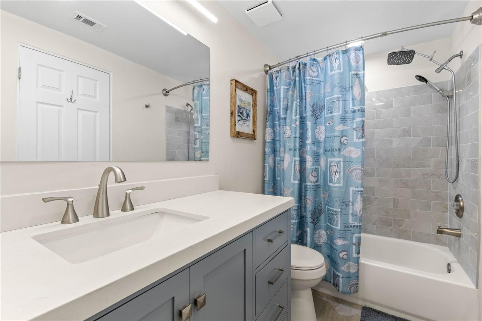 The remodeled primary bath (2025) pairs a wide gray vanity and sleek white countertop with a tiled tub/shower surround, rainfall showerhead, handheld sprayer, and updated fixtures for a clean, modern finish. - 332 WESTWINDS DR, PALM HARBOR