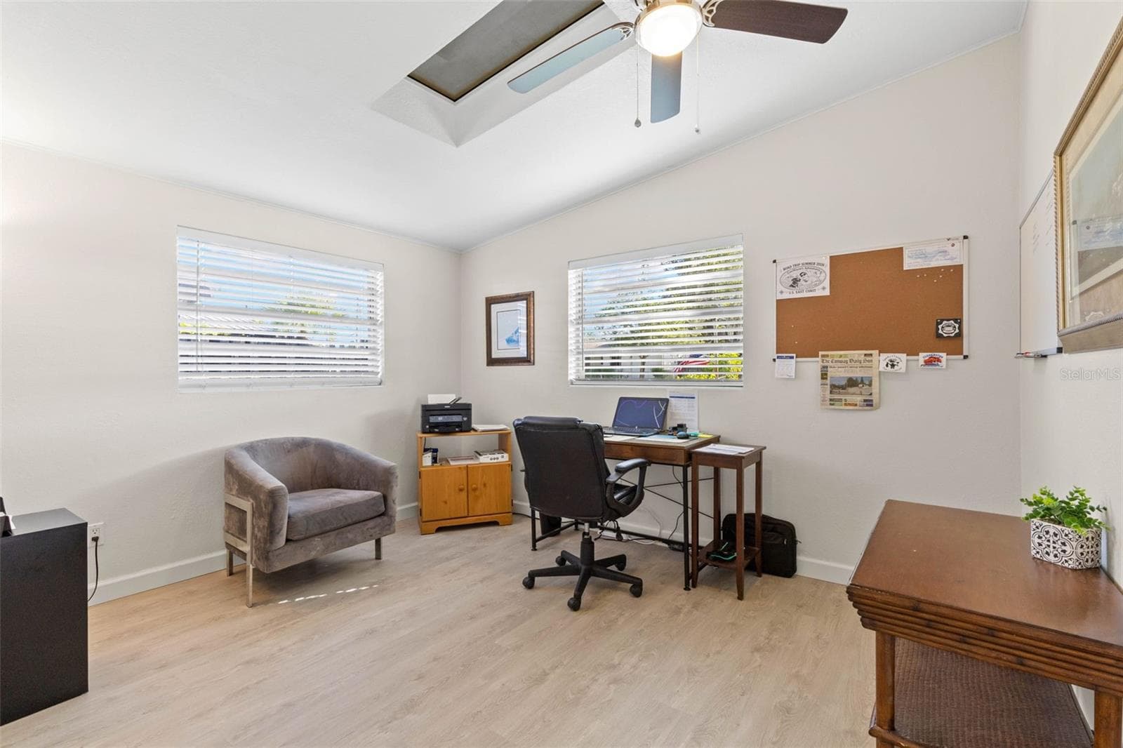 A bright secondary bedroom, currently used as a home office, offers two windows, a skylight, ceiling fan, neutral tones, and luxury vinyl plank flooring (2025), with plenty of flexibility for guests, work, or hobbies. - 332 WESTWINDS DR, PALM HARBOR